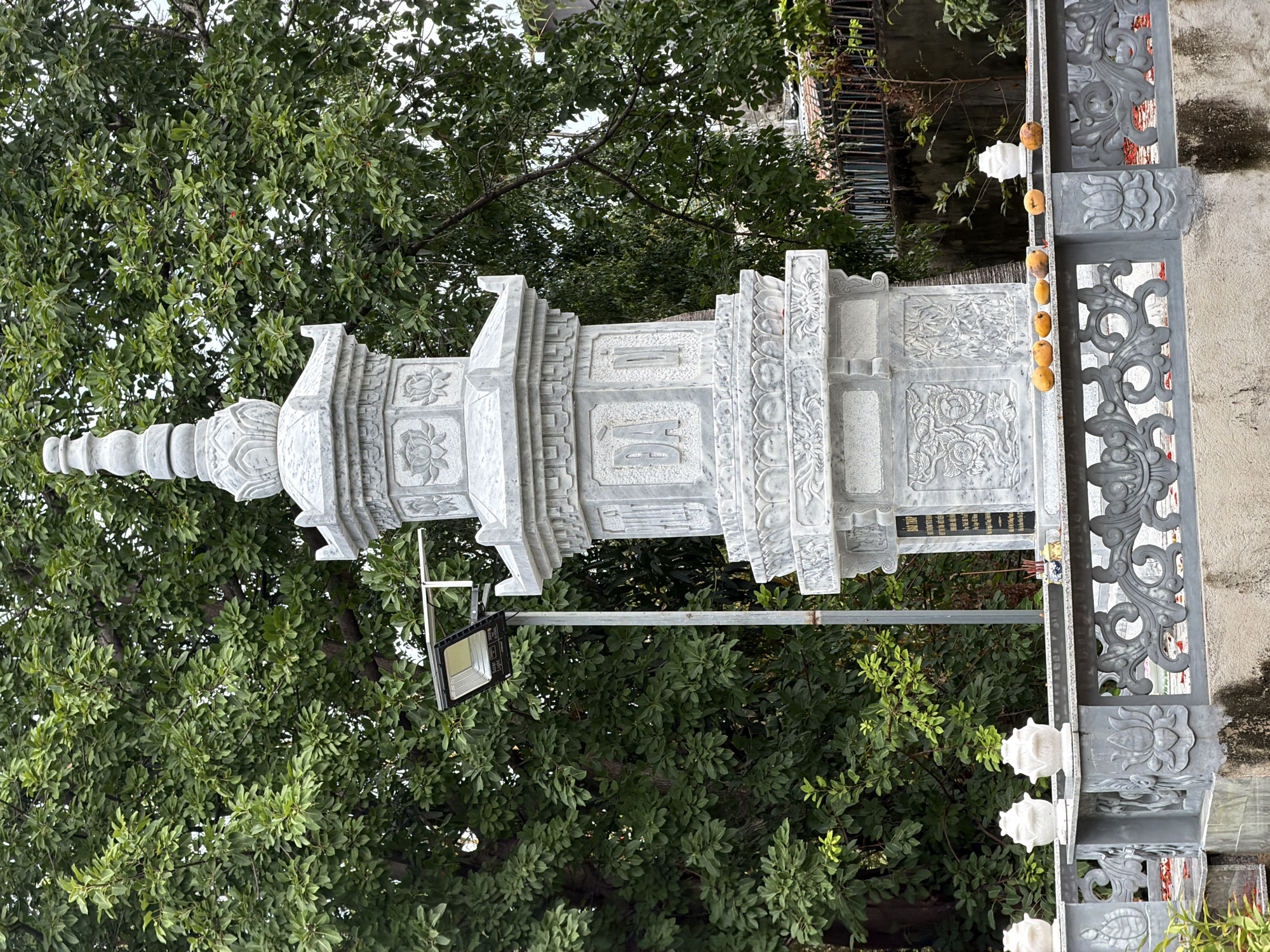 The image displays an ornate, multi-tiered stone stupa or memorial column, carved from light grey stone, set against a backdrop of lush green trees. The structure is intricately detailed with traditional East Asian motifs, including prominent lotus flower carvings. Visible Chinese characters on one central panel read "往生蓮位" (Lotus Seat for Rebirth) and "仙師仙姑" (Immortal Master, Immortal Maiden). Below these, a darker plaque contains further dedicatory text, partially legible as "佛光普照" (Buddha's light shines universally) and "先考" (deceased father), suggesting a memorial purpose.
Dense green foliage from numerous trees fills the background and much of the upper frame, indicating an outdoor, natural setting. A modern LED floodlight is mounted on a metal pole next to the stone structure. To the right, a decorative grey stone railing or gate features elaborate scrollwork and floral patterns, echoing the carvings on the central structure. A string of small, round, yellowish-orange fruits, possibly kumquats, hangs from this railing. In the far right, a textured, light-colored wall is partially visible.
The scene takes place during the daytime, under soft, diffused light, with no people present. The architectural style, carvings, and visible text point to a cultural or religious site in Cam Ranh, Vietnam, conveying an atmosphere of tranquility and cultural heritage.