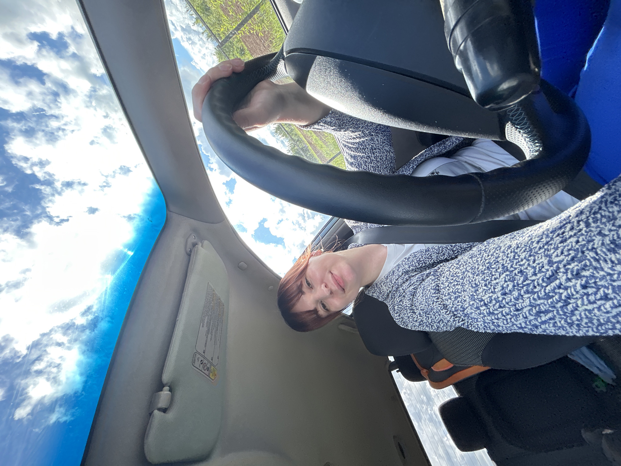 A tilted, low-angle shot captures a person in the driver's seat of a car, looking directly at the camera. The subject, a female with reddish-brown hair and bangs, wears a white t-shirt and a blue and white textured cardigan. Her hands are on the black steering wheel, which has visible stitching. Below the waist, she wears dark leggings/pants and orange and black sneakers are visible.
The scene is set inside a car, with elements of the interior such as the dashboard, a light grey sun visor with illegible warning labels, and a portion of the car roof lining visible. Through the car's windshield and side window, a bright blue sky with scattered white clouds dominates the upper and left parts of the frame, indicating a clear, sunny daytime. Beyond the car, green vegetation, possibly fields or trees, can be seen, suggesting an outdoor or rural setting. The person appears to be posing for a photograph, likely a selfie, with a neutral to slightly pleasant expression. The location context provided is Фоки, Russia.