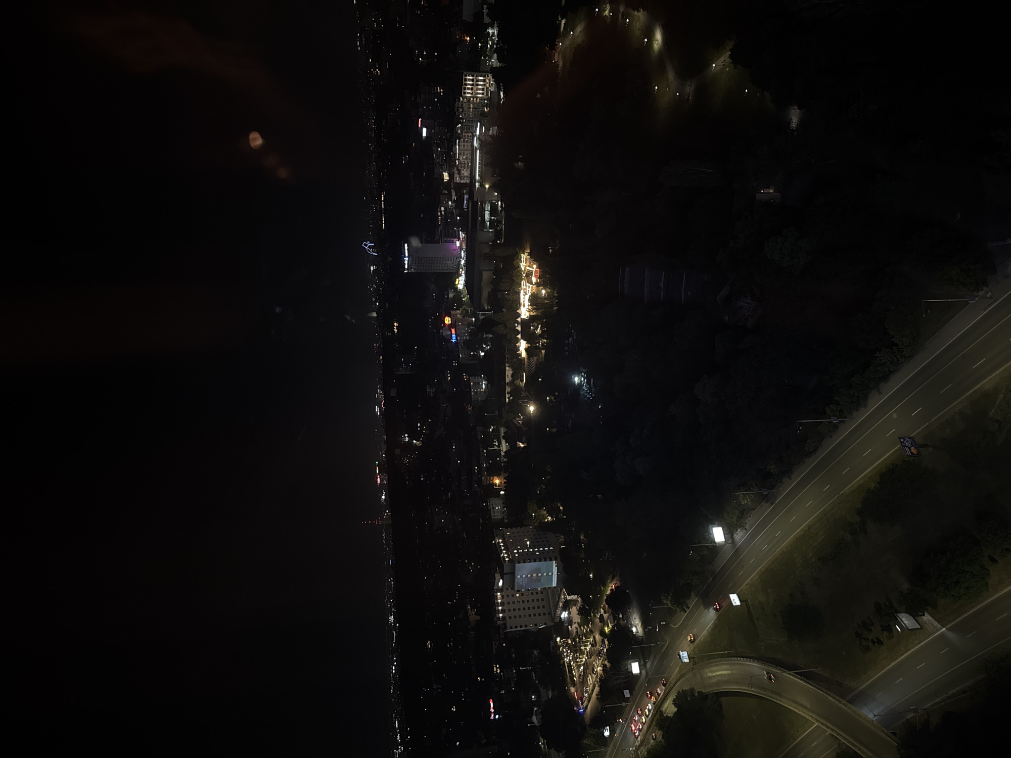 The image shows a nighttime aerial view of a city, identified as Sofia, Bulgaria, with a focus on a highway intersection and surrounding urban landscape. The darkness of the night is illuminated by numerous artificial lights from buildings, streetlights, and vehicles.
In the foreground, a multi-lane highway intersection is visible, with cars driving on the different roadways. A concrete overpass curves to the left, crossing over a lower road. Streetlights line the sides of the highways, casting pools of light. Several billboards are also present along the roadside.
Further into the scene, a large, multi-story building with many illuminated windows dominates the mid-ground. Adjacent to this building, a parking lot with many cars is visible, also illuminated by overhead lights. Beyond this complex, the city extends into the distance, characterized by a dense collection of buildings and lights. A tall, slender tower with a red light at its peak is visible in the far distance. Other structures, including what appears to be a modern office building with a blue illuminated top, are scattered throughout the cityscape. The overall environment appears to be a developed urban area with significant infrastructure and building density. The trees in the mid-ground and foreground are dark and heavily shadowed, indicating a significant amount of foliage within the city.