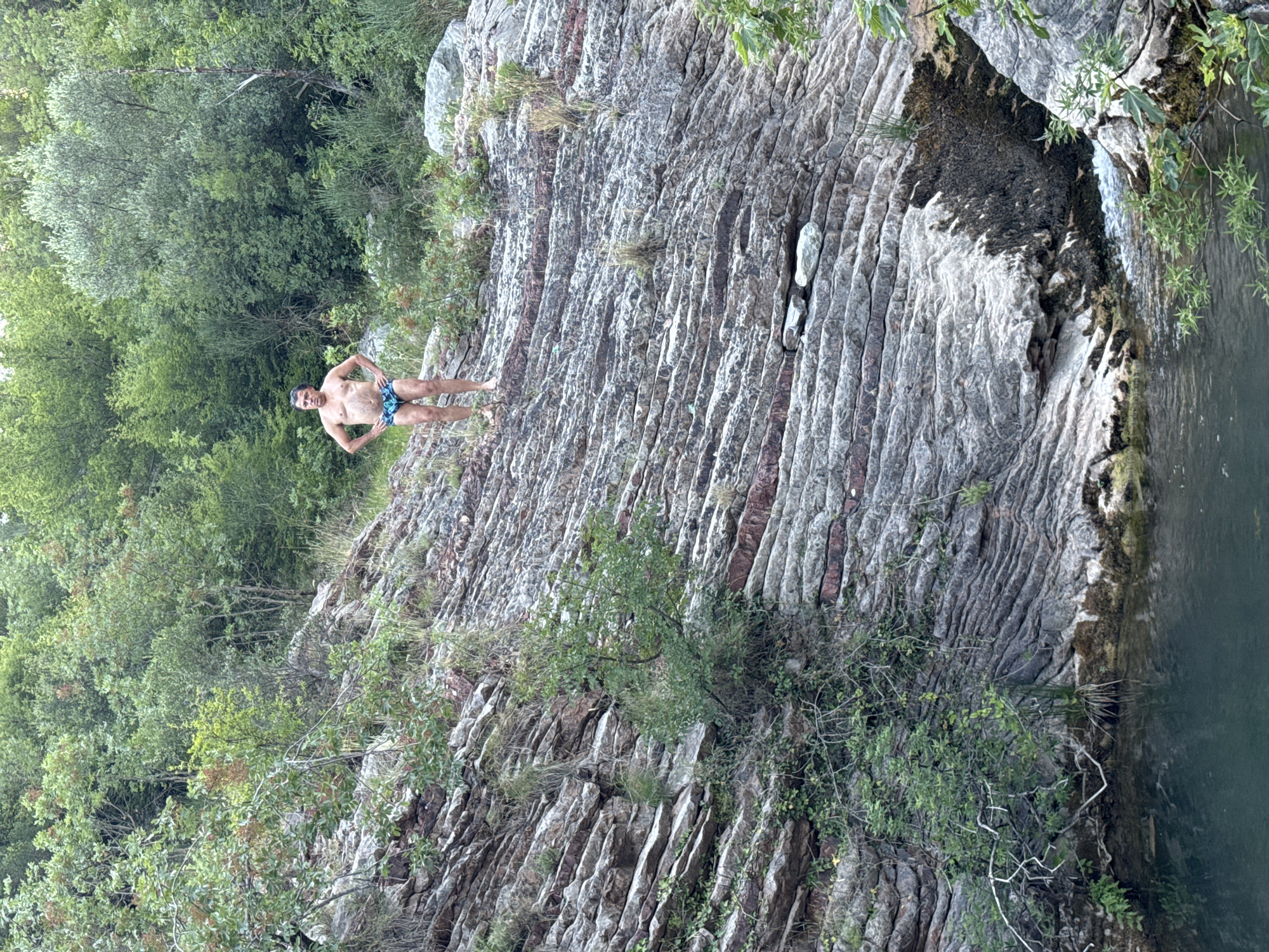 An adult male stands on a rocky incline overlooking a body of water. He is wearing blue patterned swim trunks and has his hands on his hips. The incline is composed of layered rock formations with sparse vegetation growing on it. Behind the man and the incline, a dense forest with green foliage is visible. The body of water in the foreground appears to be a natural pool or small river. The scene is set in a natural, outdoor environment, likely in the vicinity of Rafailovići, Montenegro, given the characteristic rocky terrain and lush greenery common to the coastal region.