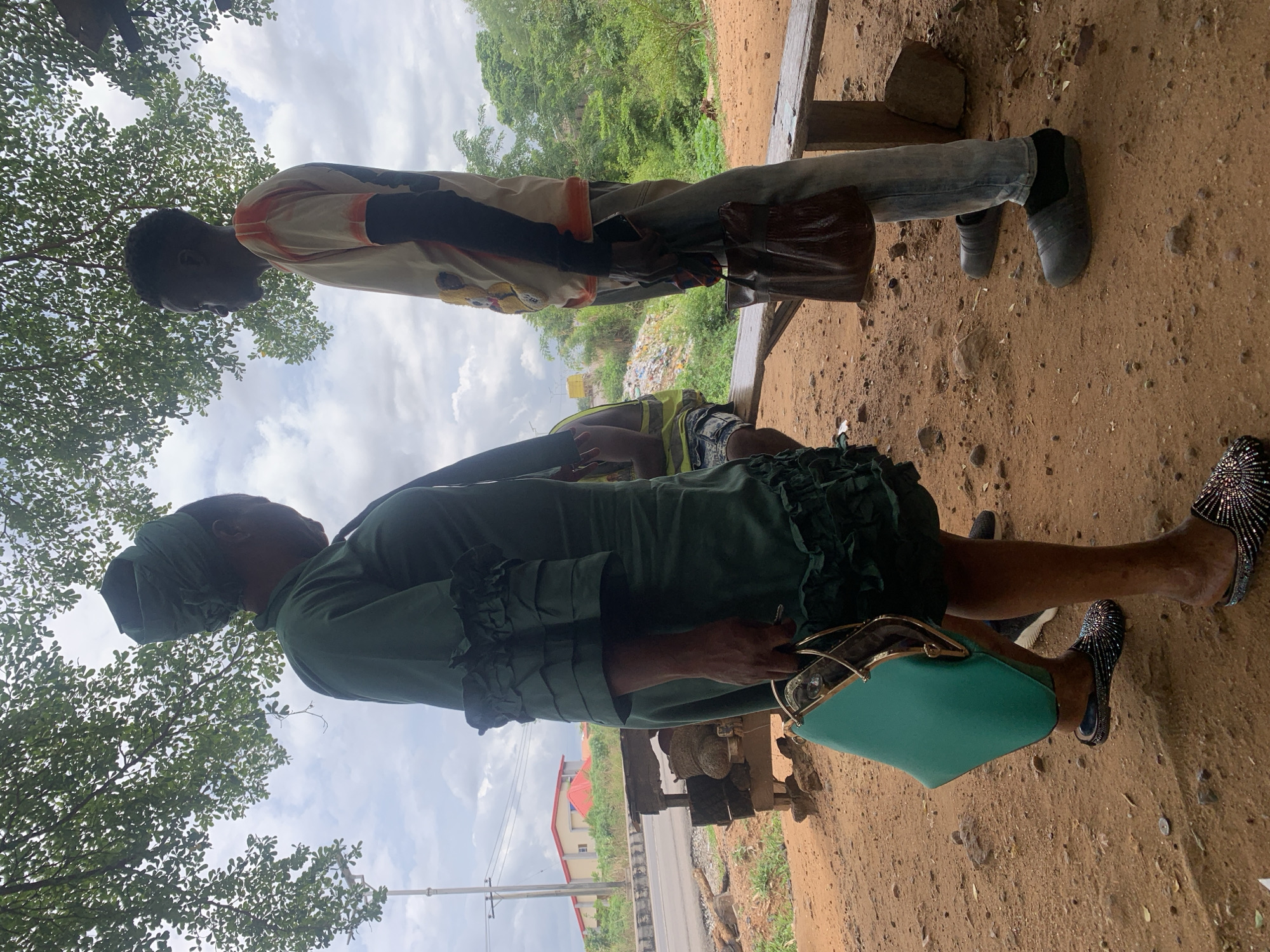 The image depicts two individuals in an outdoor setting. One individual, presumed female, wears a green dress and matching head covering. She is also wearing dark, possibly studded, shoes. She has a green handbag. The other individual, presumed male, is wearing light-colored shirt with dark and orange accents, jeans, and dark shoes. He carries a brown bag. They are near what appears to be a wooden structure, possibly a bench or a makeshift table. The ground is dusty and there is vegetation in the background. A building with a red roof is visible. The image's location could potentially be Lokoja, Nigeria.