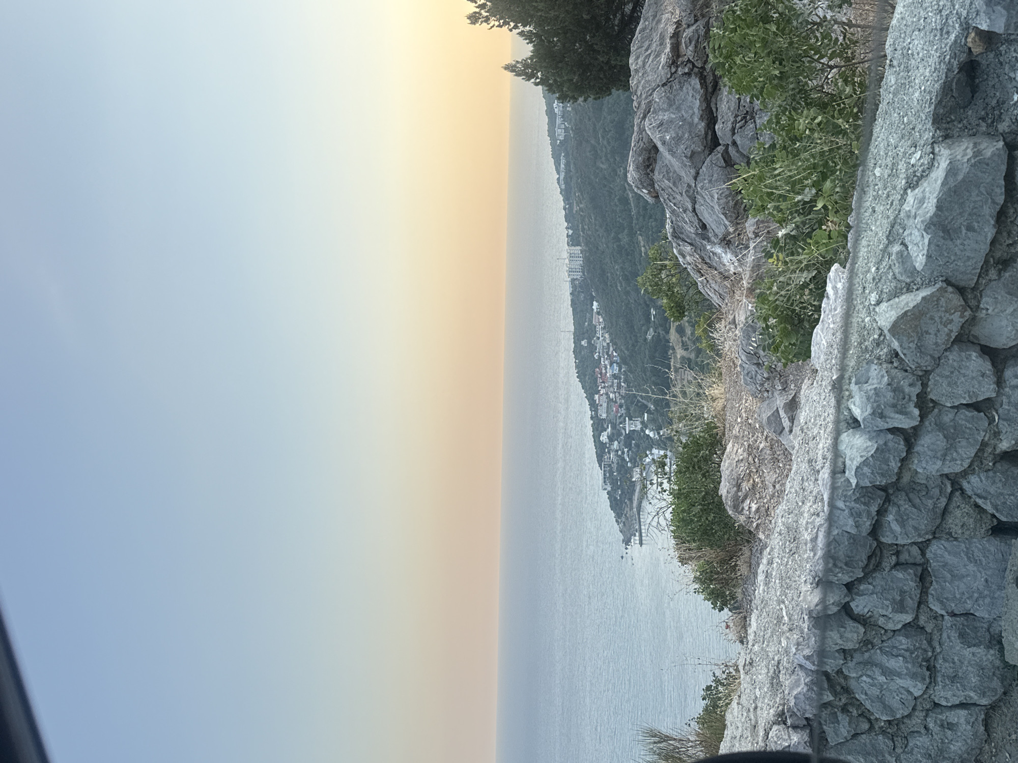 A wide shot captures a tranquil coastal scene at dusk. The sky transitions from a pale blue near the top to a soft, warm yellow at the horizon, meeting a calm, vast ocean. Along the coastline, a small town with white buildings is nestled amongst lush green hills. In the foreground, a stone wall constructed from rough, gray rocks borders the view, with some greenery growing around it. The overall impression is one of peaceful natural beauty.
