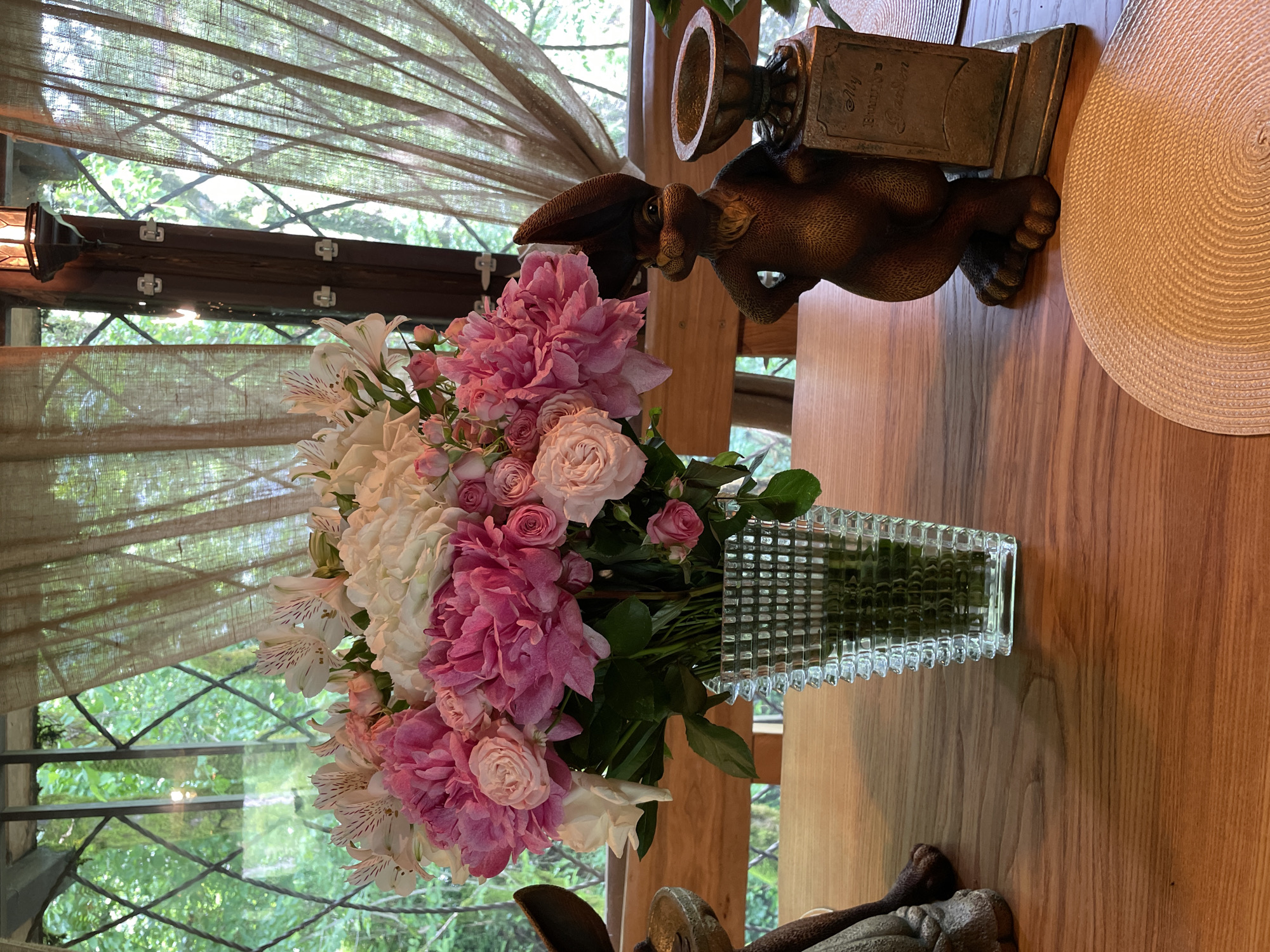 A still life photograph captures a bouquet of pink and white flowers in a clear, textured glass vase, set on a wooden table. The vase rests near a bronze rabbit figurine holding a plaque inscribed with "My Funtime Garden." A round, woven placemat partially enters the frame on the right. In the background, a window with a lattice-like design offers a view of lush greenery, possibly trees. The lighting is soft and diffused, suggesting an indoor setting with natural light filtered through curtains. The photograph has been rotated 90 degrees counterclockwise. There is no indication of this image being taken in Solokh-Aul, Russia.