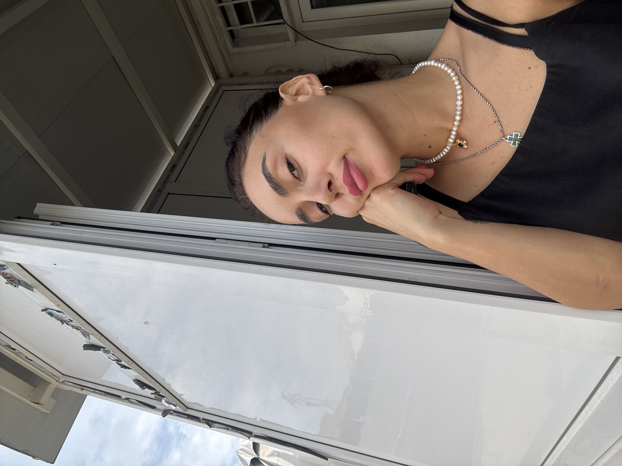A young woman with dark hair and full lips is leaning her cheek on her hand, looking at the camera. She is wearing a black tank top and a pearl necklace with a gold pendant, and another silver necklace with a blue and silver cross pendant. Her head is tilted slightly to the left, and her eyes are open. The background consists of a white structure with a window. The overall scene suggests an outdoor setting, possibly a balcony or an open doorway, with a cloudy sky visible in the upper left corner. The architecture of the building, specifically the window frames and the white panels, appears consistent with modern construction found in urban areas. While the specific location in Tbilisi, Georgia, is not definitively identifiable from the visual cues alone, the general urban environment and architectural style are plausible for this city.