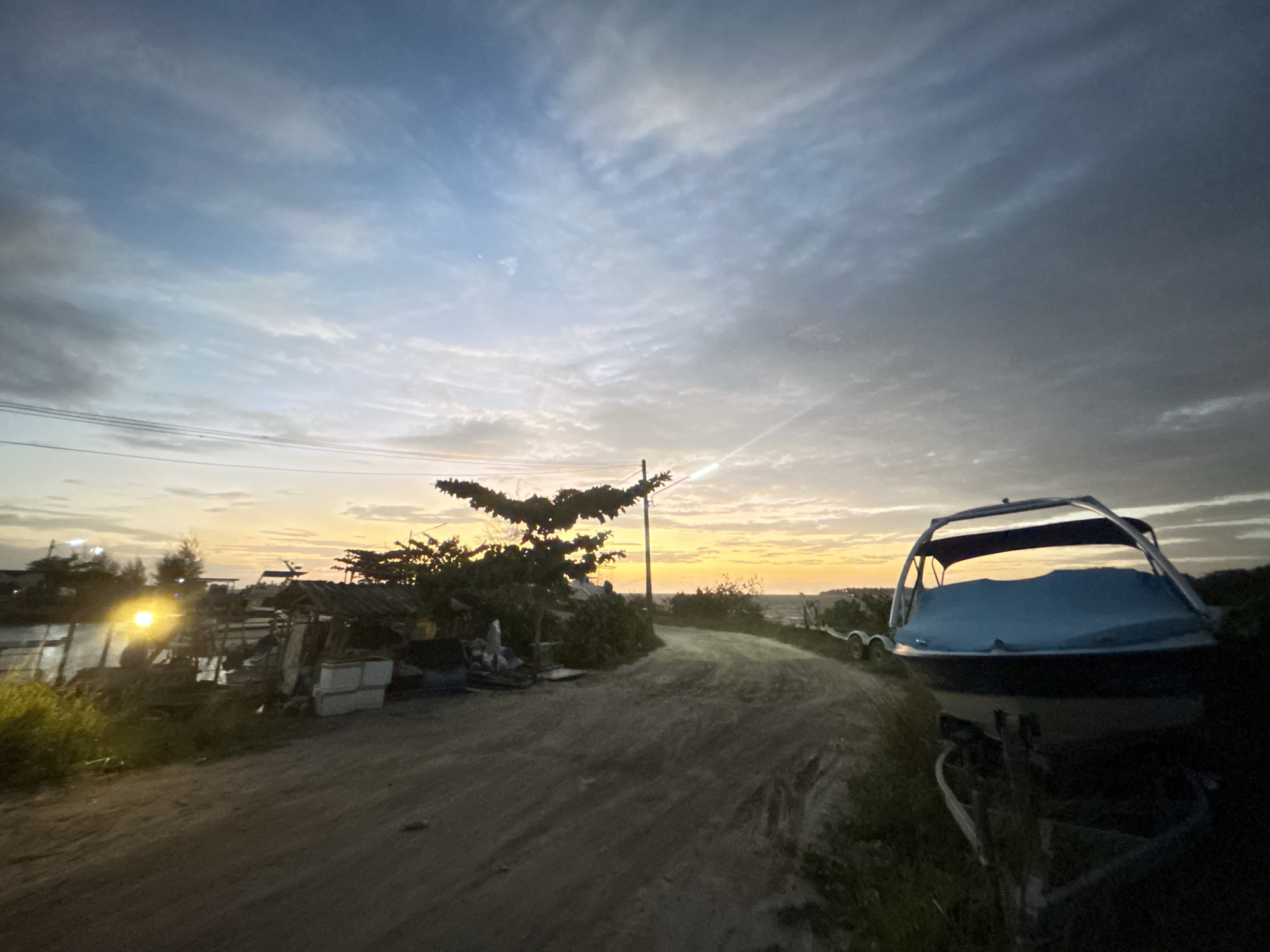 The image depicts a dirt road leading towards a body of water at dusk. The sky is a mix of blue and grey clouds, with a warm orange glow on the horizon. On the left side of the road, there is a cluster of trees and what appears to be a small structure with various objects, possibly fishing equipment or debris. A bright light source is visible on the left, illuminating the surrounding vegetation. On the right side of the road, a boat covered with a blue tarp is parked on a trailer. The overall scene suggests a rural or coastal setting in the Ko Pha-ngan District, Thailand, likely a beach or a small harbor area, characterized by a serene, quiet atmosphere as daylight fades.
