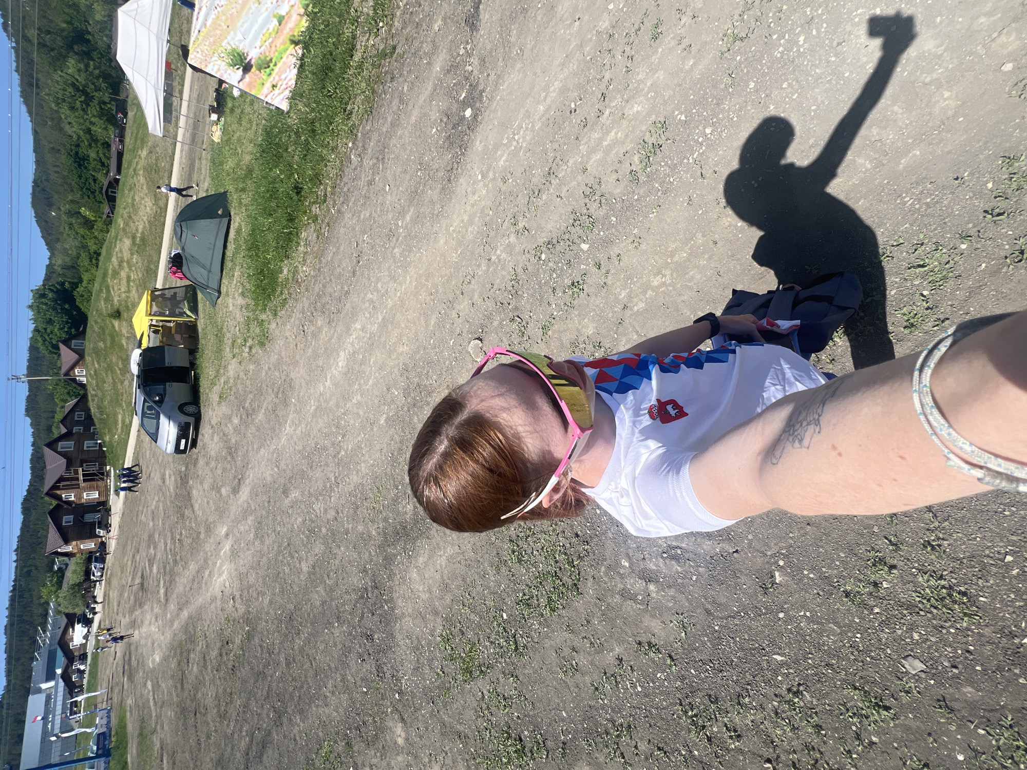 A high-angle, selfie-style photograph shows a person standing on a dirt road on a sunny day. The person has reddish-brown hair and wears pink sunglasses. They are wearing a white shirt with a design in the colors of the Slovakian flag. The person's shadow is visible on the road, holding up a camera. In the background are several tents, cars, and buildings. A hillside is also visible in the background. Based on the style and contents of the photo, it appears the person is in Khvalynsk, Russia.