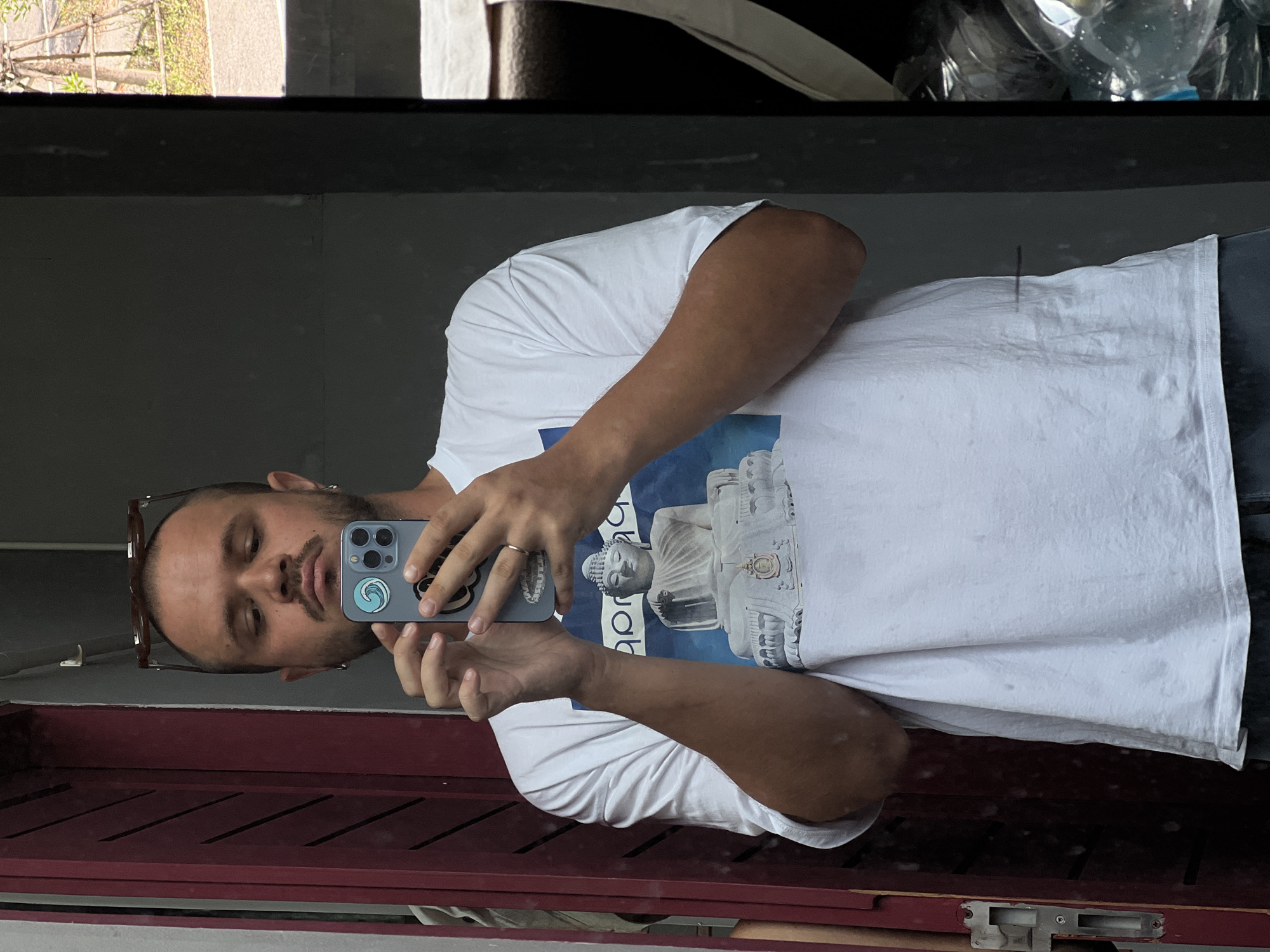 A man is reflected in a mirror, taking a selfie. He is wearing a white t-shirt with an image of a Buddha printed on it. He has short hair and a beard, and is wearing sunglasses on top of his head. He is holding a smartphone in his right hand, and his left hand is resting on his chest. The background is blurry, but appears to be an indoor or outdoor space with some greenery visible through an opening. The man is standing in front of a dark wall, with a red door on the left side of the frame. There are also some plastic bottles visible in the right side of the frame.