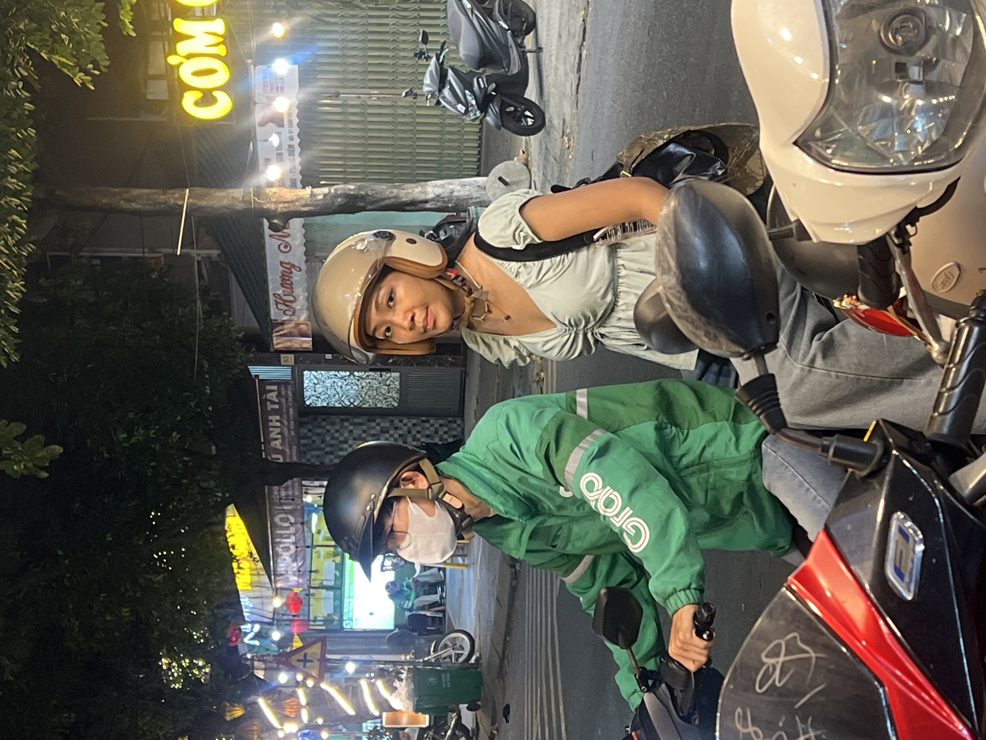 Two individuals are seated on a scooter at an intersection in Da Nang, Vietnam, at night. The person in front, wearing a green jacket with "Grab" logo, a black helmet, and a face mask, appears to be the driver. The person behind, a woman with a light-colored dress and a beige helmet, is looking towards the camera. The background features illuminated signs, trees, and other scooters. The street is paved, and there are buildings and streetlights visible.