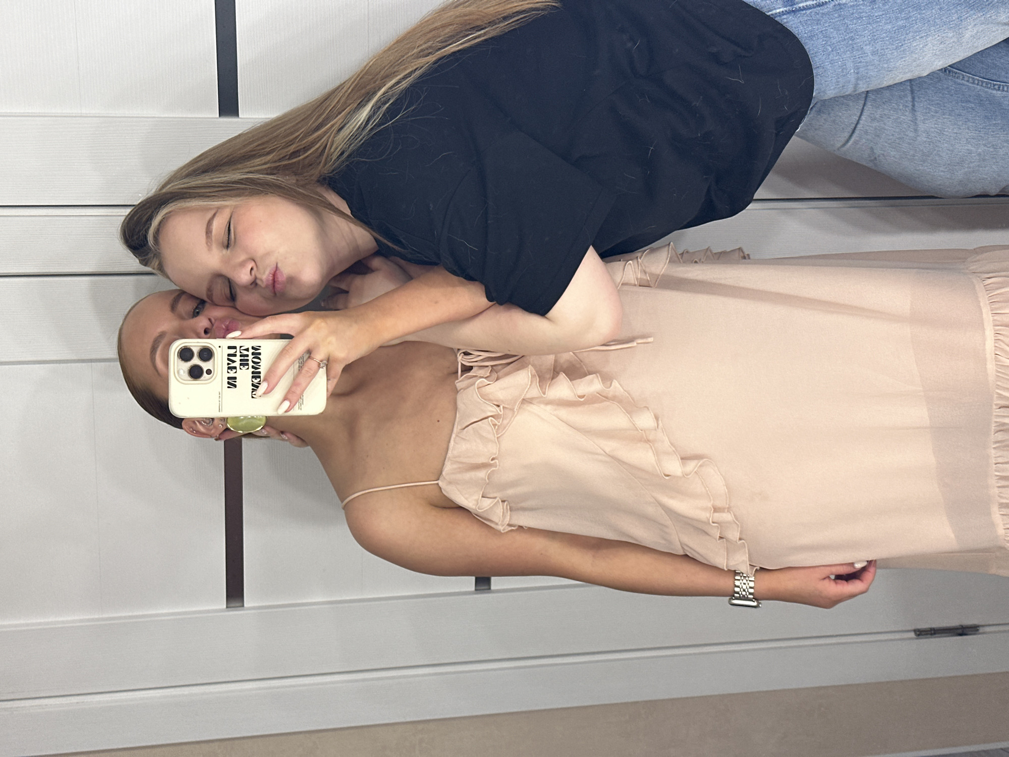 Two women are taking a selfie in front of a white paneled wall. One woman is wearing a black top and blue jeans, while the other is wearing a light pink dress with spaghetti straps. The woman in the black top is leaning towards the woman in the dress, with their faces close together as if posing for a kiss. The woman in the dress is holding a phone with a light-colored case, taking the picture. The phone case has text printed on it. The setting appears to be indoors, potentially a dressing room or a house. I cannot verify the exact location within the city of Chaikovsky, Russia based on the visual information.