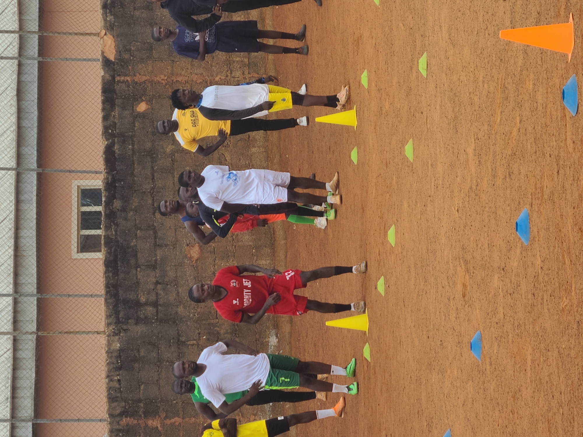 The image depicts a group of soccer players lined up in two rows on a dirt field in Unknown, Unknown. They appear to be doing drills, indicated by the various colored cones placed on the ground. The players are wearing different colored athletic wear: yellow, white, red, green, and blue. Some players have their hands on their hips. A brick wall borders one side of the field with a caged window.