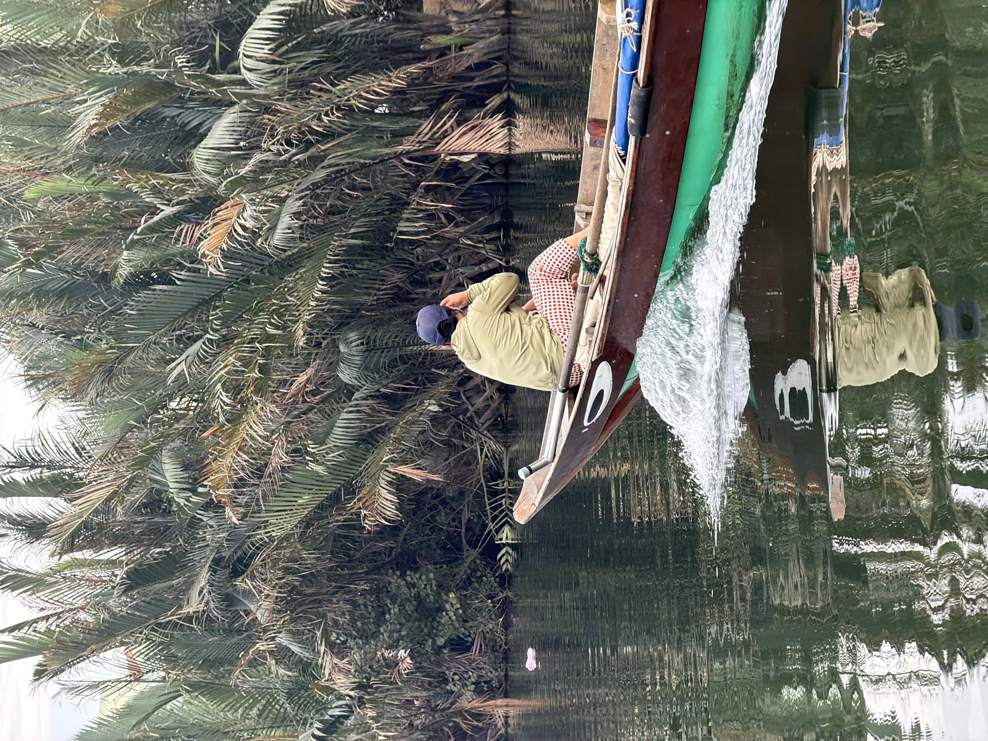 The image captures a tranquil river scene in Hội An, Vietnam, featuring a person in a traditional wooden boat navigating a narrow waterway. The subject, seen from behind, is wearing a light-colored long-sleeved shirt, checkered shorts (red and white or light brown and white), and a dark blue baseball cap. He appears to be either operating or riding in the boat.
The boat is long and slender, made of wood with a distinctive green stripe running along its side. A white wake is visible at the boat's side, indicating it is actively moving through the water. The bow of the boat features a decorative eye-like carving. Blue elements, possibly ropes or coverings, are also visible along the boat's edge.
The waterway is bordered by dense, lush foliage, dominated by numerous palm-like trees, characteristic of tropical or subtropical environments like the nipa palms found in Vietnam. The surface of the water clearly reflects the surrounding greenery and parts of the boat, suggesting relatively calm conditions with ripples created by the boat's movement. The lighting suggests it is daytime, possibly with diffused sunlight, creating a soft and peaceful ambiance. There is no visible text in the image.