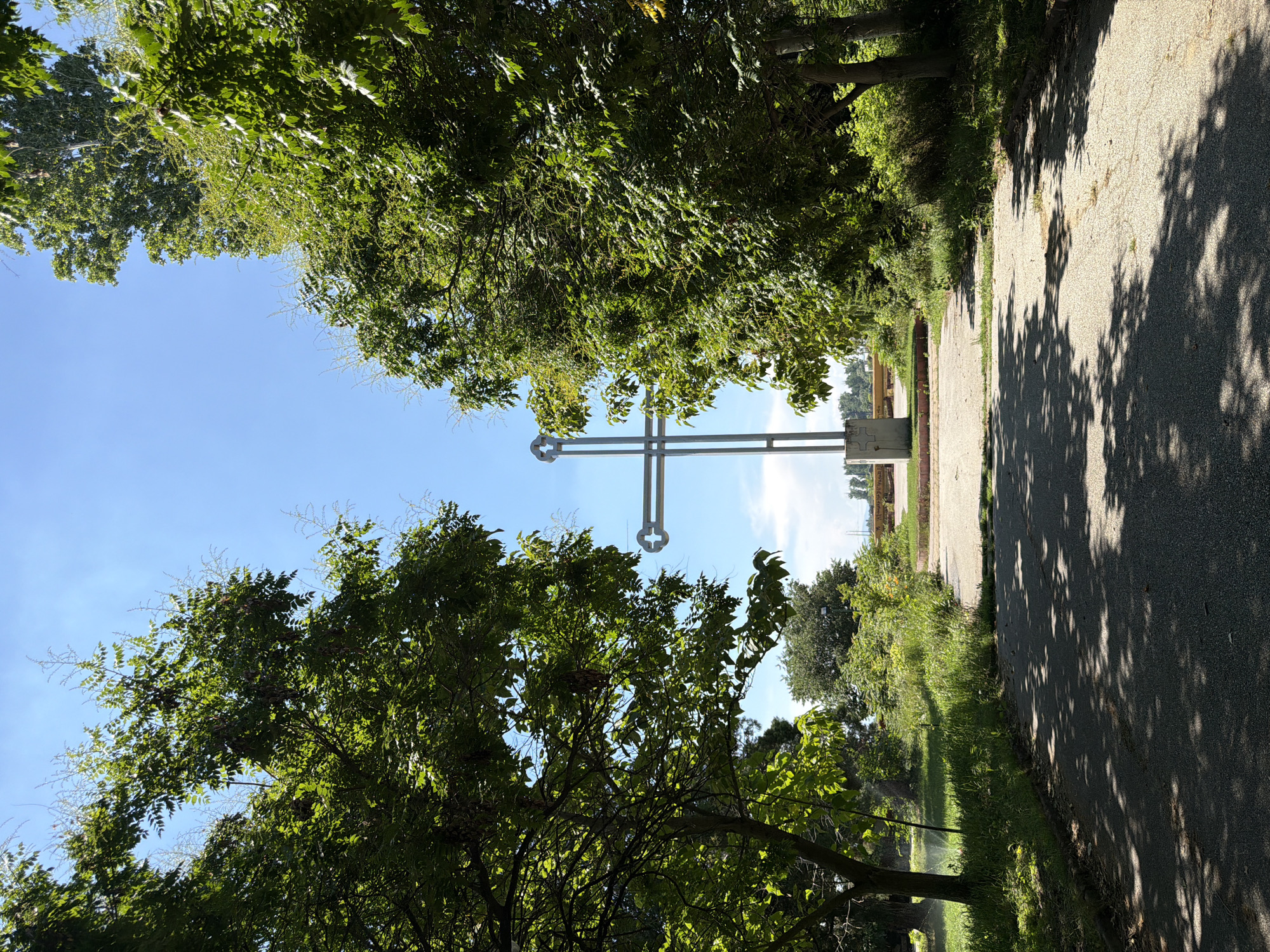 The image shows a large, modern, silver-colored cross standing in an open area surrounded by lush green trees. The cross has a hollow circular design at its intersection and is supported by a concrete pedestal. A paved pathway leads towards the cross, with shadows of tree branches dappled across it. The background reveals a bright blue sky with a few wispy clouds. The vegetation is dense, with trees and bushes creating a natural, park-like setting. The scene appears to be a serene outdoor space, possibly a memorial or a religious site. The location is identified as the Saint George Chapel in Pazardzhik, Bulgaria.