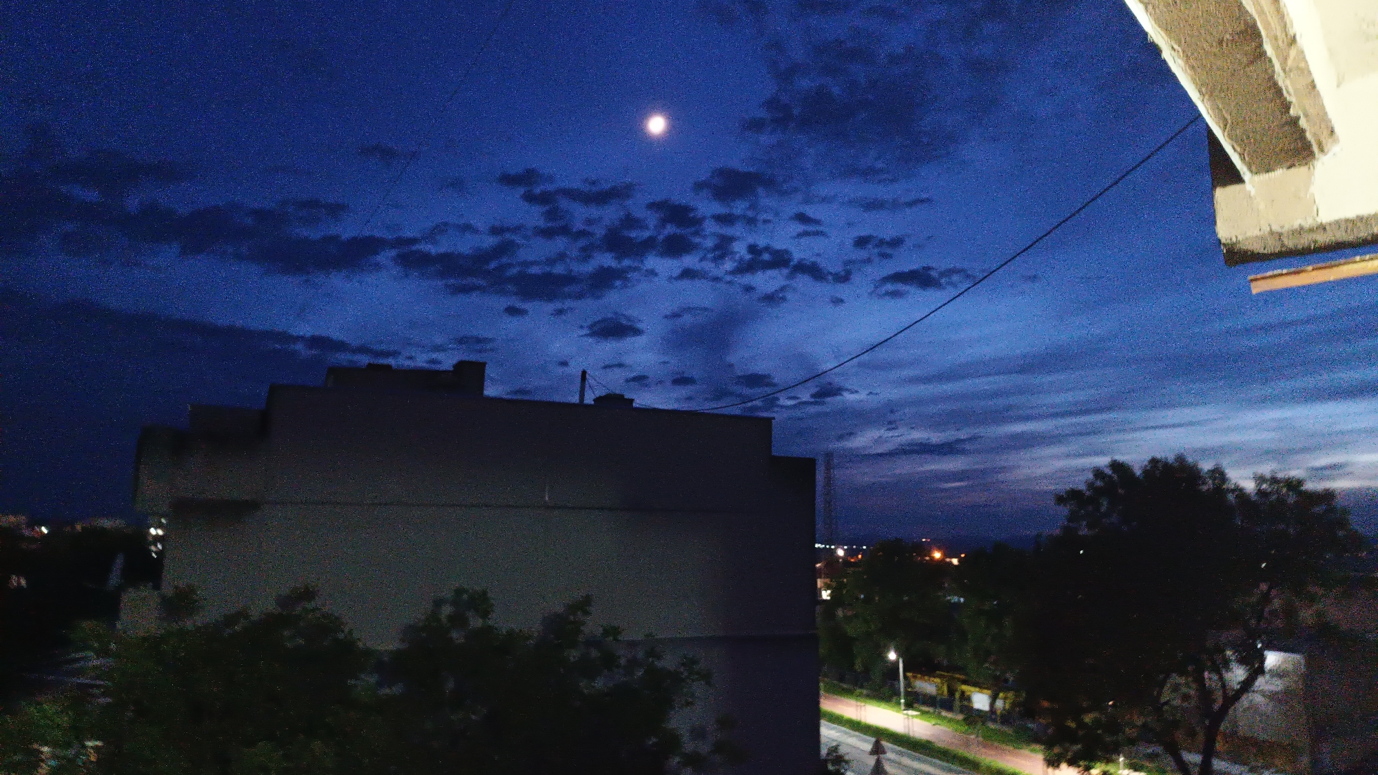 A nighttime cityscape is captured from an elevated perspective, likely a balcony or high window. The sky is a deep blue with scattered clouds and the moon prominently visible. A large building dominates the left side of the frame, while trees line the bottom edge. A road with a street light runs along the right side, and distant city lights are visible on the horizon.