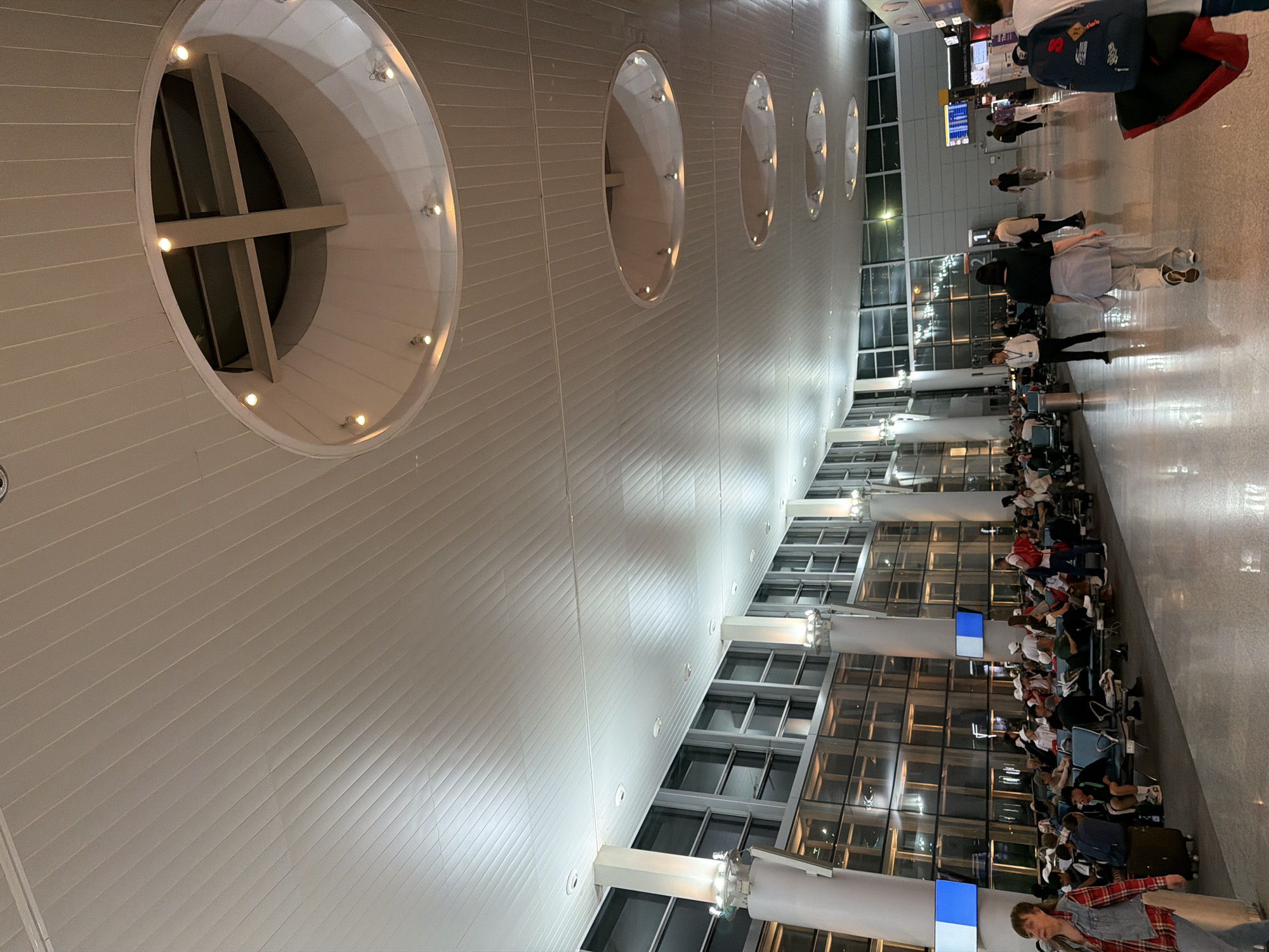 This image shows an interior view of Zvartnots International Airport in Yerevan, Armenia. The shot is taken from an angle looking up at the ceiling, which features a series of large, circular architectural details with inset lighting. The ceiling is composed of linear panels.
Below, rows of chairs are positioned in front of large windows. People are seated and standing in the waiting area. Some are walking. A flight information display system is visible in the background. Luggage is visible near the seated people. The floor is polished.