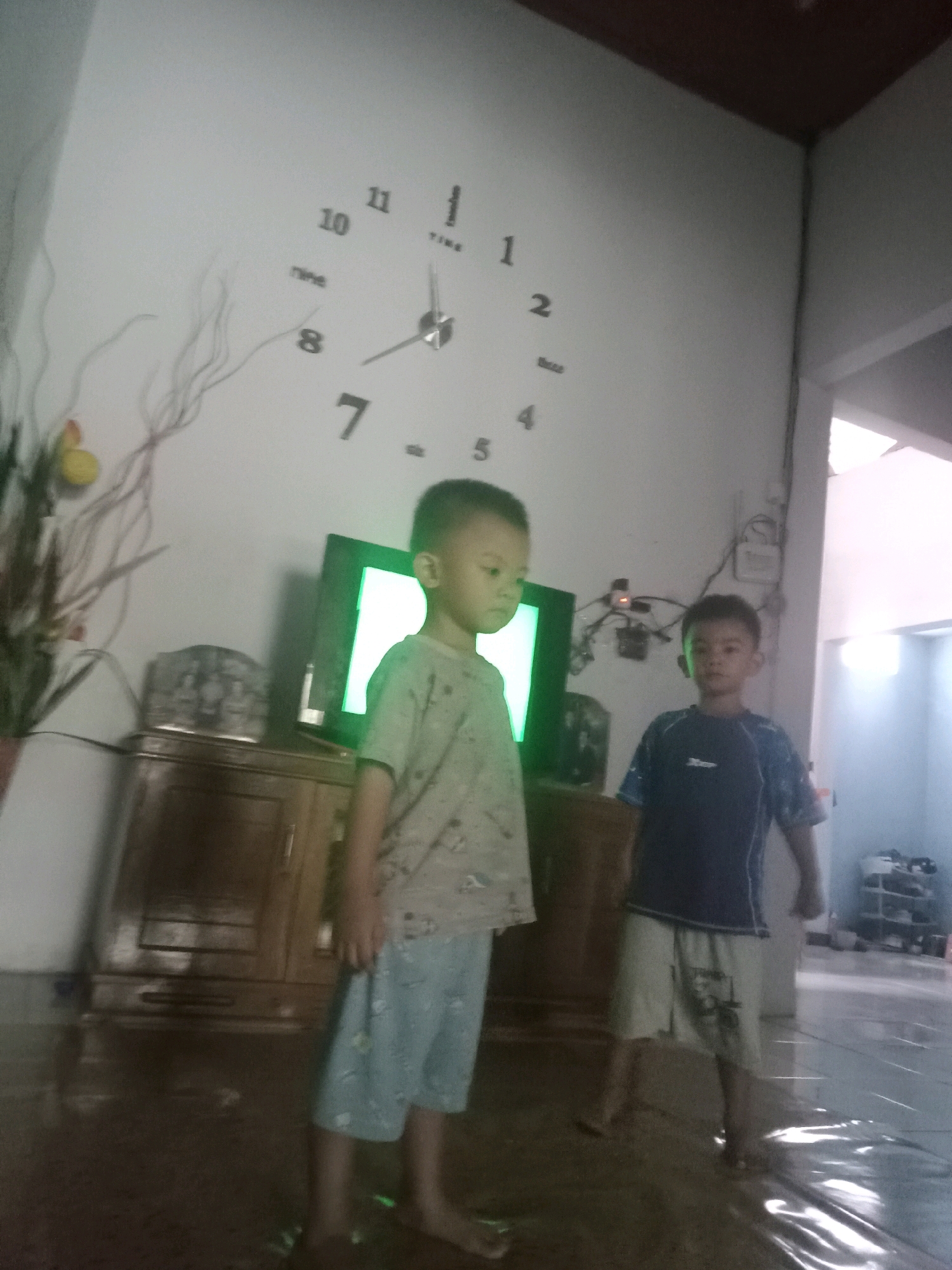 The image depicts an indoor scene featuring two young boys standing on a reflective surface. The boy on the left is wearing a light-colored t-shirt with a print and blue shorts. The boy on the right is wearing a dark blue t-shirt with a white stripe on the sleeve and light-colored shorts. Behind the boys, a television screen displays a bright, possibly green, light. A wooden cabinet is visible beneath the television. On the wall in the background, a large, modern clock with black numerals is prominently displayed, showing the time to be approximately 7:10. A decorative element resembling branches is also present on the wall. The overall environment suggests a residential setting. The image's quality is blurry, indicating it was likely taken with a low-resolution camera or in low lighting conditions. No specific landmarks or details definitively place the scene in Baubau, Indonesia, beyond the provided prompt.