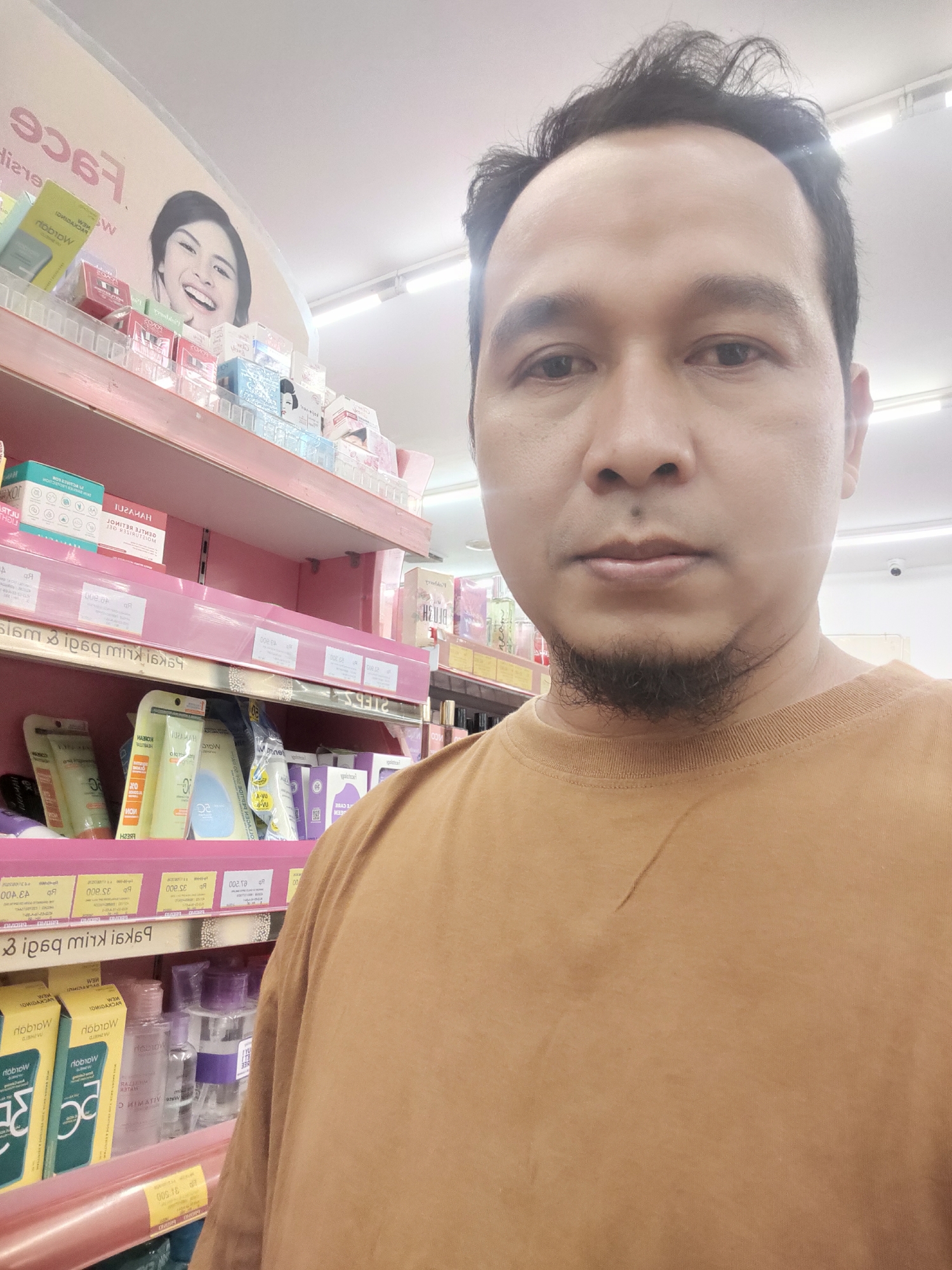 The image is a selfie taken inside a store, likely a pharmacy or cosmetic shop, in Desa Banyumas, Indonesia. The foreground is dominated by the face of a man with dark hair and a goatee, looking directly at the camera. He is wearing an orange t-shirt. Behind him, shelves are stocked with various beauty products, including boxes of skincare items and bottles. A poster featuring a smiling woman is visible on the top shelf. The lighting suggests indoor fluorescent lighting.