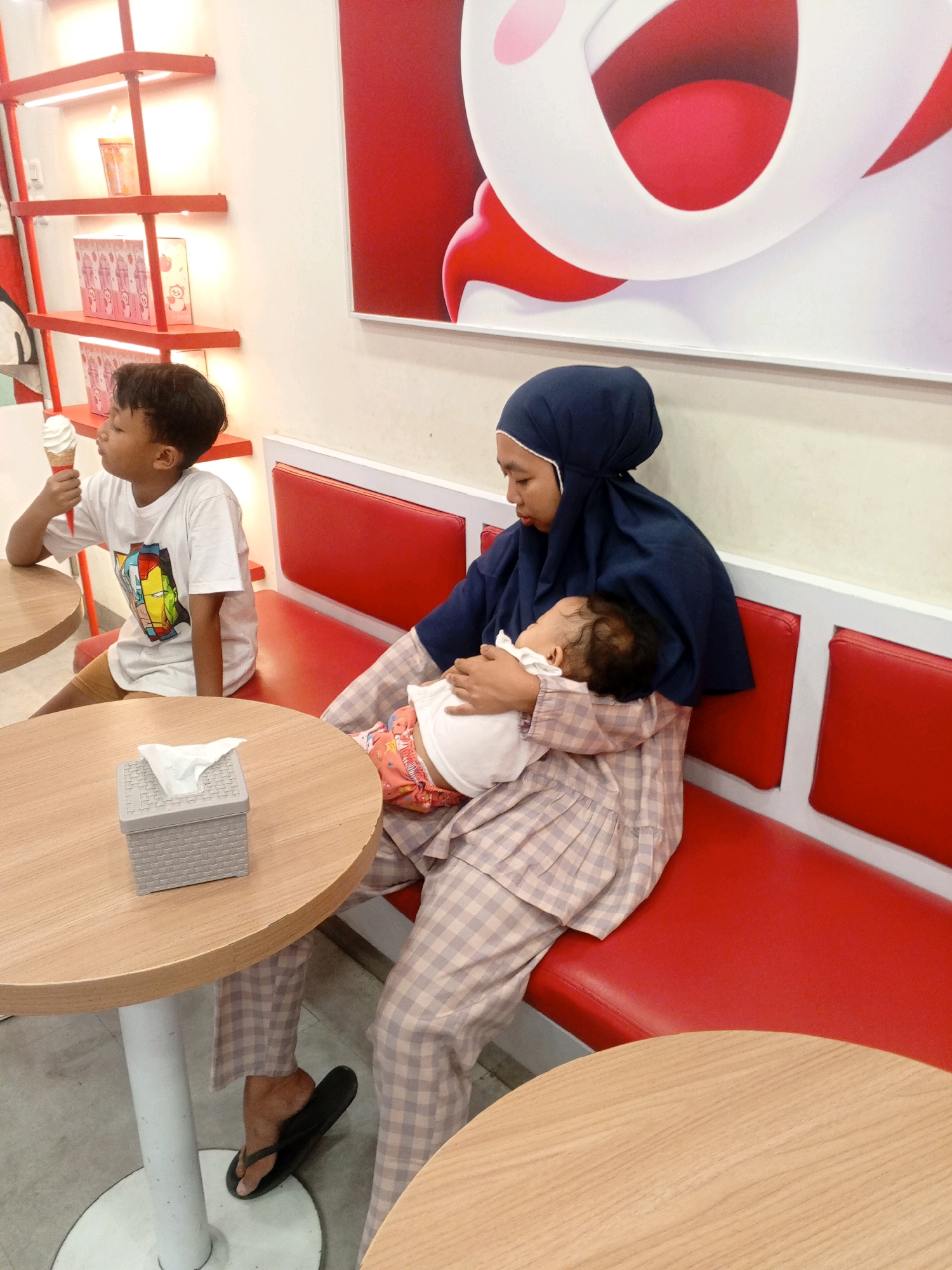 A young boy is sitting at a round wooden table in a brightly lit establishment, holding an ice cream cone and looking to his left. He is wearing a white t-shirt with a graphic design and light brown shorts. Beside him, a woman wearing a navy blue hijab and a plaid dress is holding a sleeping infant. The infant is wearing a white onesie and colorful patterned bottoms. They are seated on a red booth with white trim. A tissue box is on the table between the boy and the woman. In the background, shelves with various items are illuminated by bright lights. The setting appears to be a restaurant or ice cream parlor in Gabus, Indonesia.
