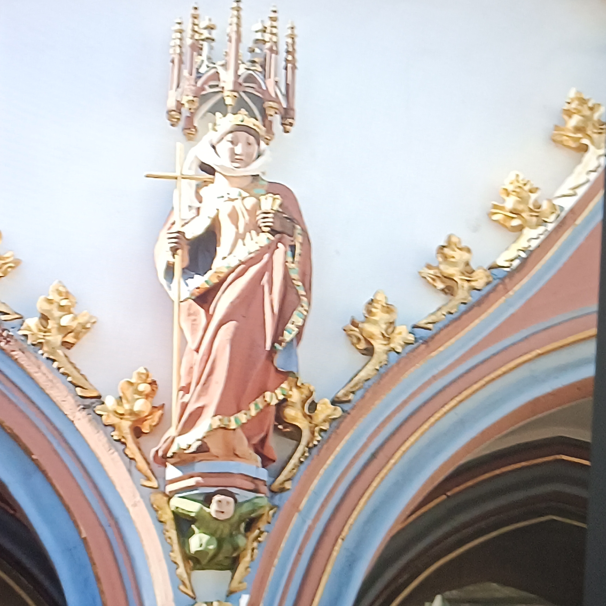 The image shows a statue of a robed figure wearing a crown, holding a cross and a book. The statue is part of the facade of a building in Rüsselsheim am Main, Germany. The building's facade is decorated with ornate architectural elements, including gothic-style spires and gold embellishments. The statue itself appears to be made of painted wood or stone. The figure's attire includes a pinkish-brown robe with a decorative trim, and a white veil or head covering. Below the statue, there is a smaller, green carved figure with a face. The architectural details of the building feature colorful arches painted in shades of blue, pink, and gold. The overall style suggests a historical or religious building, possibly a church or a town hall. The lighting in the image indicates it was taken during the daytime, with sunlight casting shadows.