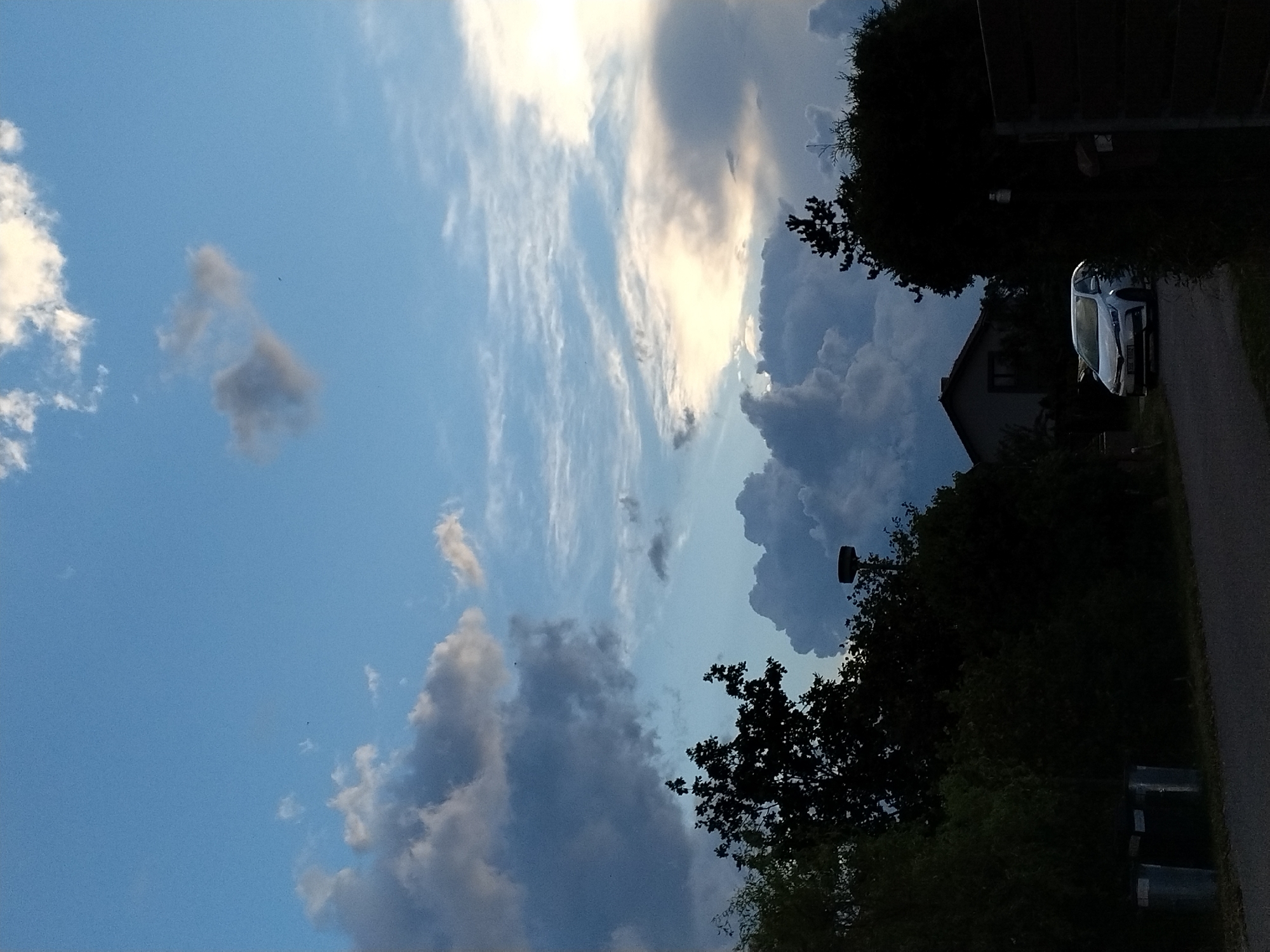 A white sedan is parked on a paved road in the rural village of Dobev, Czechia, with dense green foliage and trees lining the left side of the road. A modern house with a dark roof is visible behind the trees on the left. The sky above is a mix of blue with dramatic, dark clouds and lighter, wispy clouds, suggesting a dynamic weather condition. A street light is visible above the treeline on the right side of the image. Two grey trash bins are positioned near the edge of the road on the left. The overall lighting suggests it is either early morning or late afternoon.