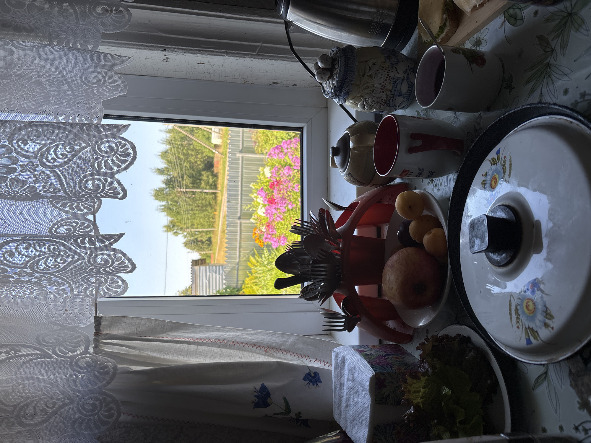 The image depicts a kitchen window view in Дунилово, Russia, overlooking a garden with pink and yellow flowers. Inside, a tablecloth covers a table laden with breakfast items. A stack of napkins sits on the left, next to a plate of lettuce. A red utensil holder overflows with cutlery. A bowl contains an apple and some small fruits. Two mugs, one red and white, the other white with a strawberry design, sit on the table, one filled with a dark beverage. A small teapot and a decorative jar with a lid shaped like fruit are also present. To the right, a silver electric kettle is visible. A partially eaten sandwich with cheese and greens rests on a wooden board. A large pot lid with a floral design is in the foreground, reflecting the overhead light. Lace curtains frame the window.