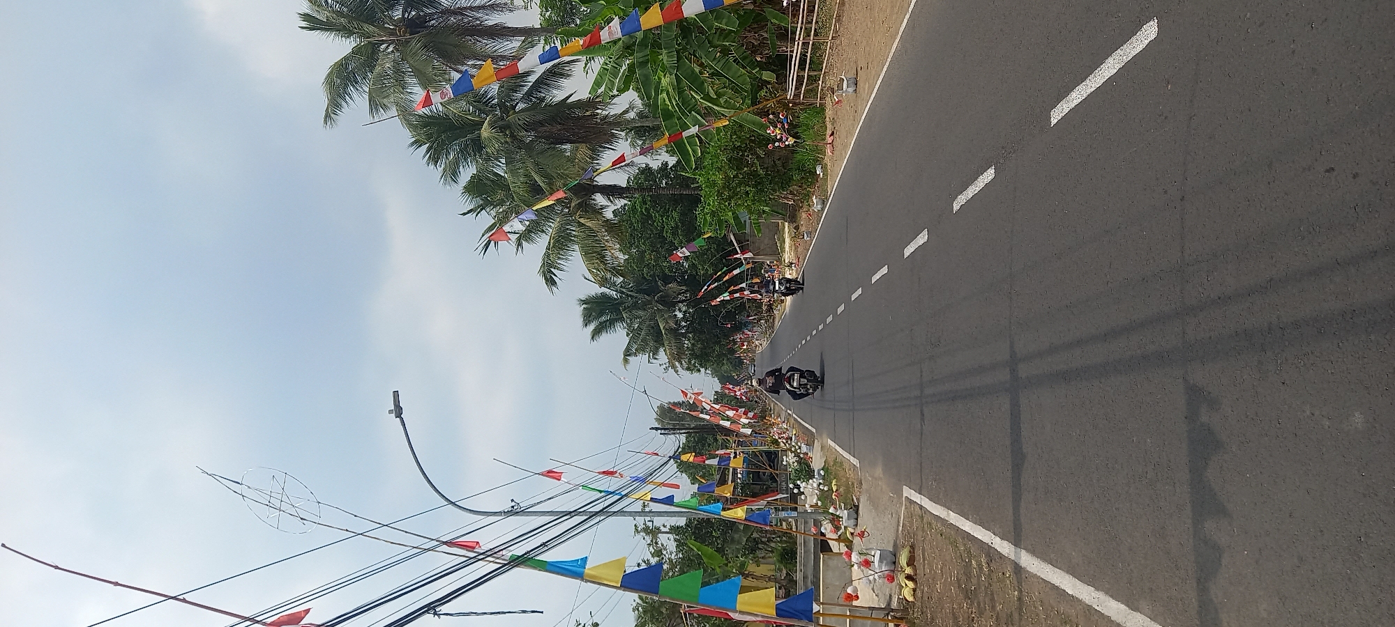 The image depicts a road in the city of Banjar, Indonesia, lined with colorful triangular flags and palm trees. The road is asphalt with white dashed lines marking the lanes. Two motorcycles are visible on the road, one in the foreground with a rider wearing a dark helmet and shirt, and another further down the road. The sky is overcast. The flags are of various colors, including red, white, blue, yellow, and green, and are strung along bamboo poles. Some flags appear to be in the Indonesian national colors of red and white. Vegetation, including palm trees and leafy green trees, lines the sides of the road. Electrical wires are also visible, strung between poles. The overall scene suggests a festive or celebratory atmosphere, possibly related to a local event or holiday.