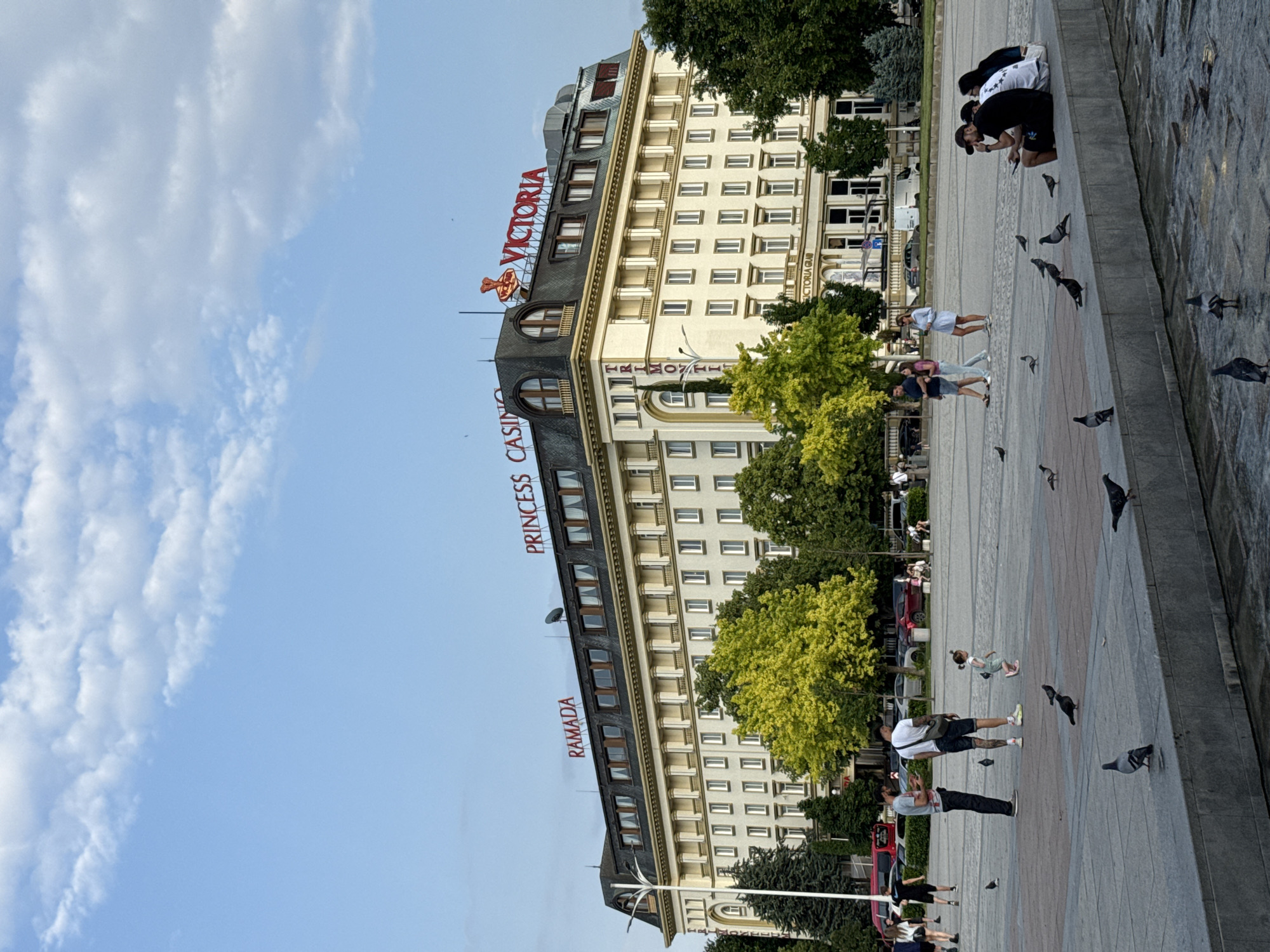 The image captures a public square in Plovdiv, Bulgaria, featuring the Grand Hotel Plovdiv (also known as Trimontium Hotel, Princess Hotel, or Ramada Plovdiv Trimontium). The multi-story building occupies the mid-ground, characterized by a light-colored facade with numerous rectangular windows and a prominent dark grey mansard roof. Red illuminated signs on the roof spell "RAMADA" on the left, "PRINCESS CASINO" towards the center-right, and "VICTORIA" with a crown-like emblem on the far right. The word "TRIMONTIUM" is visible vertically on the left corner of the building. Green deciduous and coniferous trees partially obscure the ground level of the hotel.
In the foreground, a paved plaza extends towards the viewer. A raised concrete or stone barrier, possibly part of a fountain or planter, spans the lower portion of the image, with shallow water visible in the basin below it.
Several individuals are present on the plaza. On the left, two adult males stand, one in a grey t-shirt and dark trousers, the other in a white t-shirt and dark shorts with a cross-body bag. A young child, possibly a girl, in a light-colored garment and pink shoes, walks towards the right between them and the seated group. On the right, four individuals are seated on the concrete barrier, one wearing a dark t-shirt and a cap. Additional people are visible walking in the background.
Numerous pigeons are scattered across the plaza and on the concrete barrier, with some also present in the shallow water. The sky is light blue with scattered white cumulus clouds, particularly dense on the right side. The scene is illuminated by daylight.