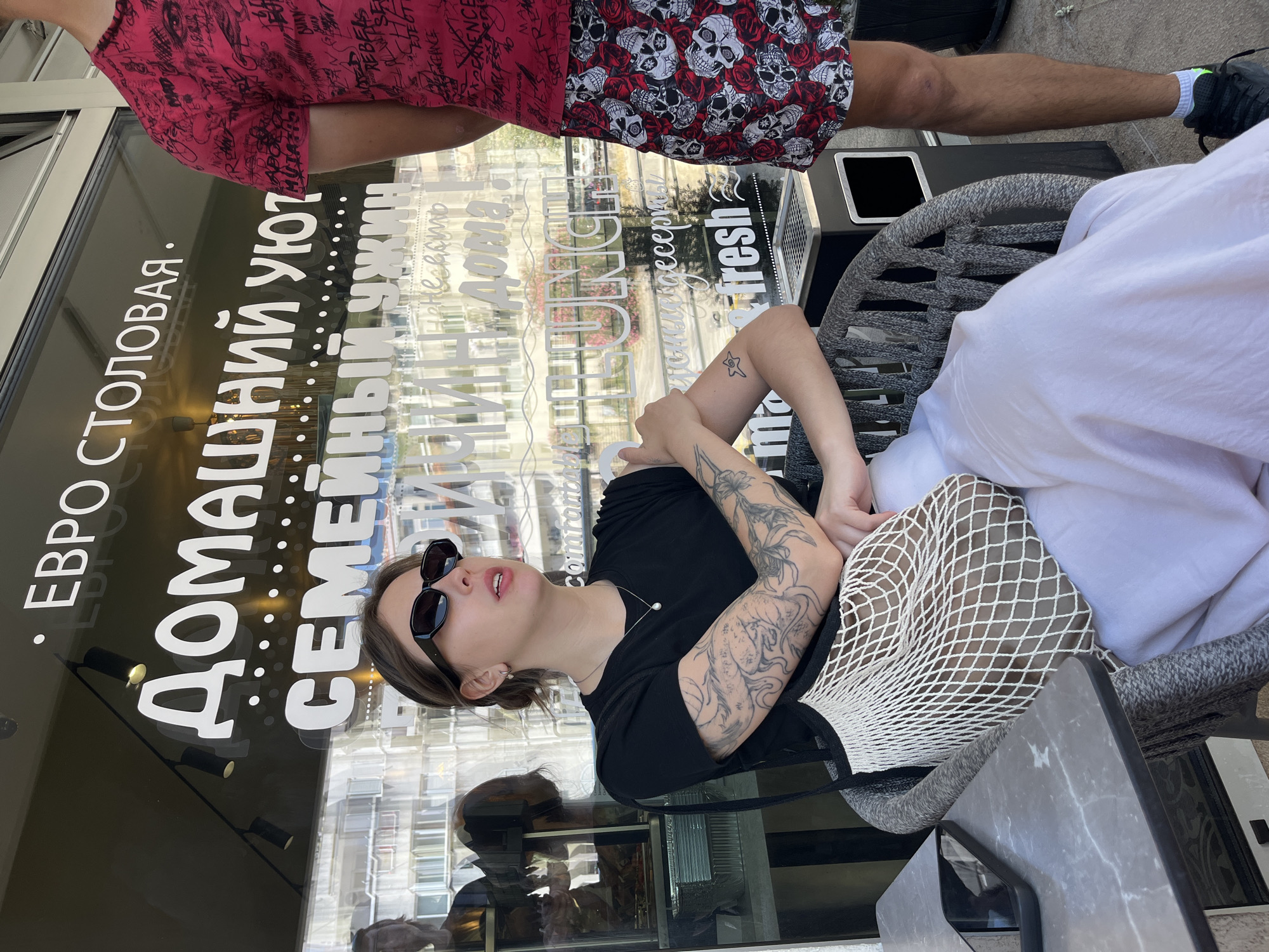 The scene is an outdoor cafe or restaurant in Donji Pobori, Montenegro. A young woman is seated in a grey woven chair at a grey marbled table, facing slightly right and looking upwards. She wears black rectangular sunglasses, a black short-sleeved t-shirt, white wide-leg pants, and a silver necklace with a single pearl pendant. Her arms are extensively tattooed, with a large, intricate design covering her left forearm and upper arm, and a smaller star-like tattoo on her right forearm. A white mesh net bag rests on her lap. To her right, a man stands partially visible from the chest down. He wears a red t-shirt with black text and designs, and red shorts patterned with white skulls and red roses. He has black athletic shoes with green accents and white socks, and a visible scuff on his right knee. A black outdoor bin or ashtray with a metal grate top is positioned between the woman's chair and the man's leg. Behind them is a large glass storefront window displaying white Cyrillic text, including "ЕВРО СТОЛОВАЯ" (EURO CANTEEN/DINING ROOM), "ДОМАШНИЙ УЮТ" (HOME COMFORT), and "СЕМЕЙНЫЙ УЖИН" (FAMILY DINNER), along with English words like "fresh" and "LUNCH". The window reflects surrounding multi-story buildings and hints of greenery under bright daylight conditions.