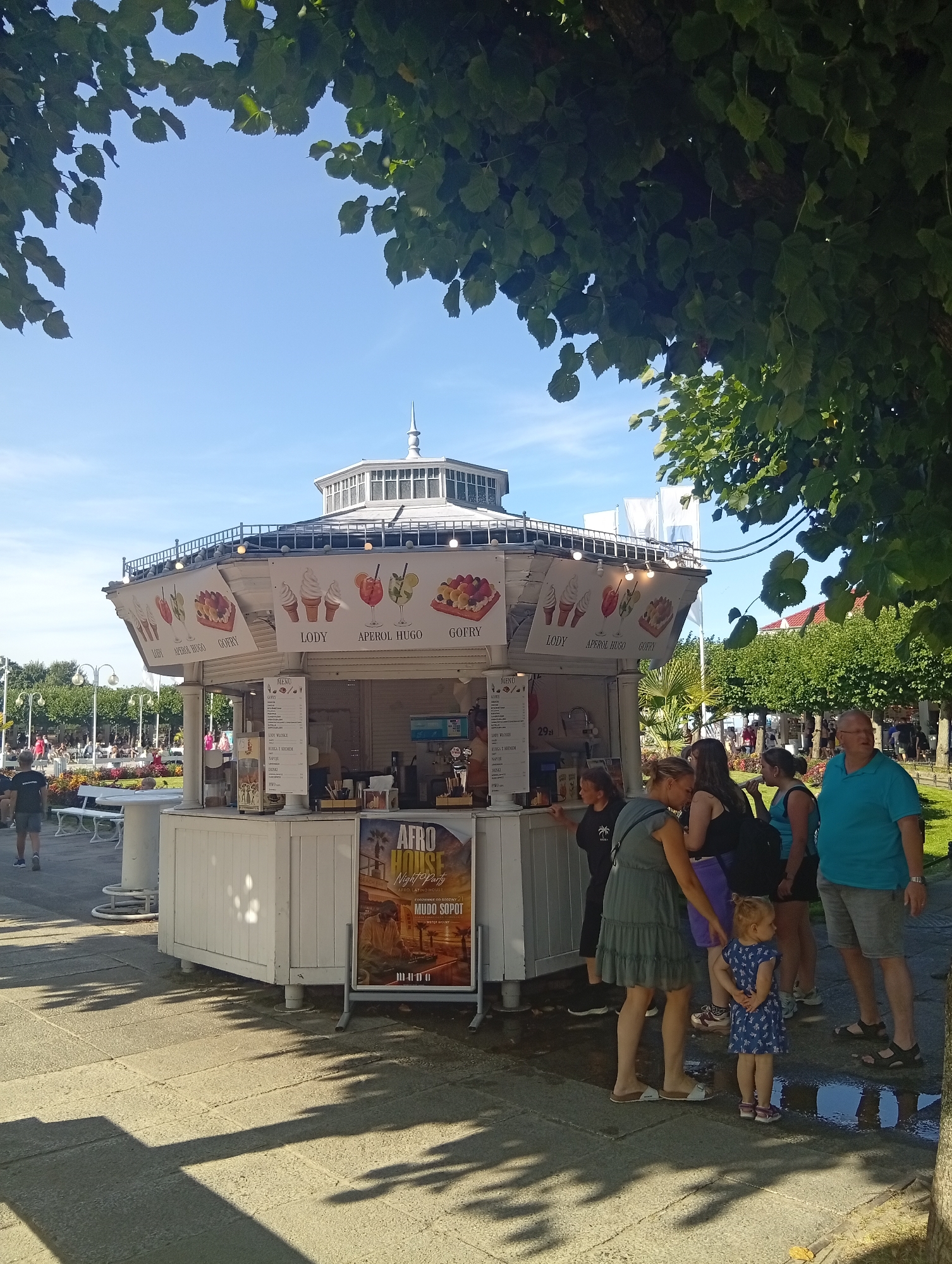 The image depicts a sunny outdoor scene in Sopot, Poland, likely along the waterfront or a busy pedestrian area. In the foreground, a white, octagonal kiosk with a decorative roof and railings is prominently featured. The kiosk displays advertisements for "LODY" (ice cream), "APEROL HUGO," and "GOFRY" (waffles), suggesting it is a food and drink establishment. A menu is visible on a stand in front of the kiosk.
Several people are gathered around the kiosk. A group of women and a young child are standing near the service window, appearing to interact with staff or decide on orders. To the right, a man in a blue shirt and shorts stands observing the scene. In the background, more people are visible walking and sitting on benches amidst trees and lampposts, indicating a public space. A sign for "AFRO HOUSE NIGHT PARTY" with "MUBO SOPOT" is posted on the side of the kiosk. The ground is paved with stone tiles, and shadows from trees are cast across the area. The sky is clear and blue.