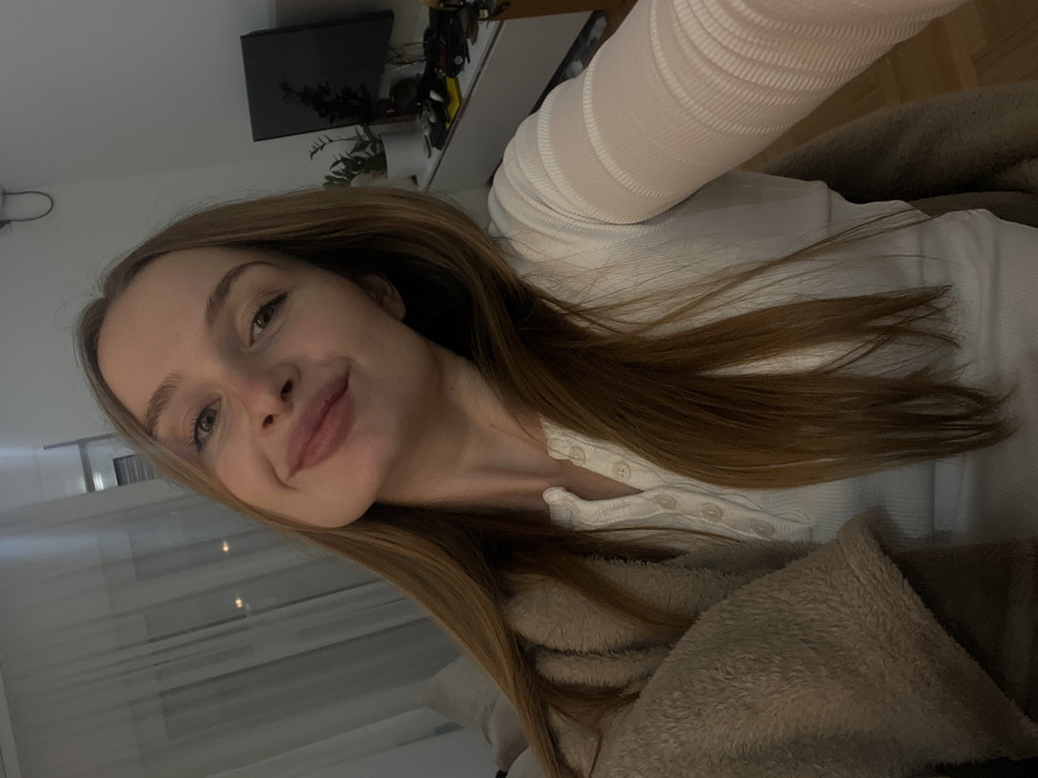 A young woman smiles gently towards the camera, displaying light brown hair parted to the side. She wears a white, long-sleeved shirt with small buttons at the neckline. A cozy, light brown blanket or jacket surrounds her shoulders. Behind her, a window with sheer white curtains is visible. In the background, a television sits atop a light-colored cabinet, adorned with a plant. The image has a casual, relaxed aesthetic.