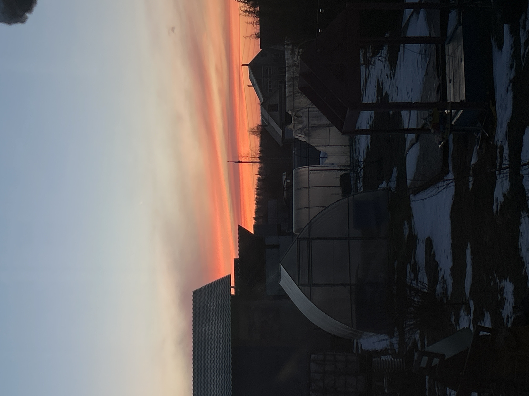 The image shows a sunset in Ольховка, Russia. The sky is a gradient of orange, pink, and blue. In the foreground, there are several greenhouses and a building with a corrugated metal roof. Some snow is on the ground. The scene is dark due to it being a sunset.