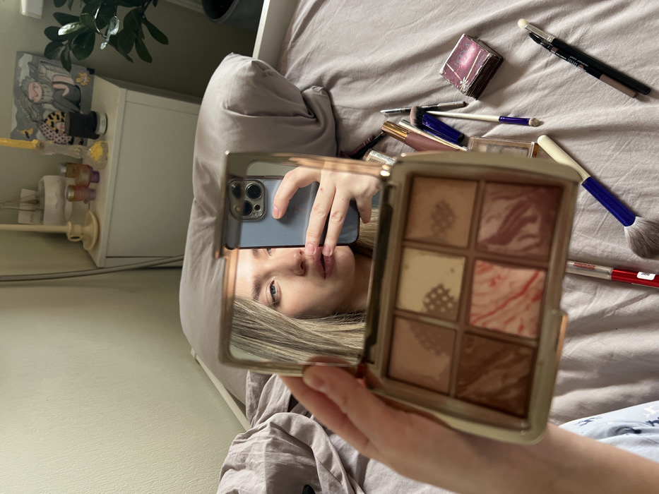 The image displays an indoor scene within a bedroom located in Saint Petersburg, Russia. A fair-skinned female with light hair is partially visible through her reflection in the mirror of an open, gold-toned makeup palette. Her left eye, nose, and lips are clearly reflected, while her right hand, holding a light blue smartphone with a visible triple camera array, obscures a portion of her face in the reflection. Her left hand holds the makeup palette towards the viewer. The palette contains six pans of makeup powders in various shades of brown, beige, and pink. Some pans feature grid-like patterns, indicative of product texture or previous application, while one pan displays a distinct marbled pink and white pattern.
The immediate foreground and midground feature crumpled light gray bedsheets and a matching pillow. Scattered on the bed to the right of the palette are various makeup items, including several brushes (one large with a blue handle), lip gloss tubes, an eyeliner pencil, and a small dark-red compact labeled "ROSE". In the background, a white nightstand is visible, topped with a light-colored candle holder containing a yellow candle, several small bottles (possibly perfumes), and a small framed artwork depicting abstract figures. A green potted plant is partially visible in the far right background, against a light-colored wall. The individual is using the smartphone to photograph her reflection in the makeup palette's mirror.