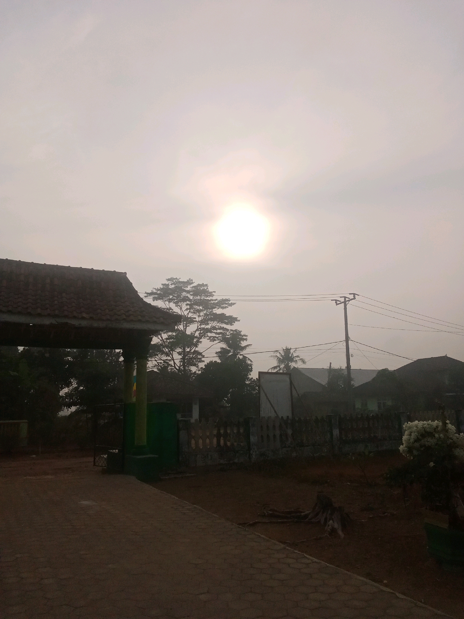 The image depicts an outdoor scene at what appears to be dawn or dusk, with a hazy sky and the sun emitting a soft glow. In the foreground, a paved walkway leads towards the left side of the frame, where a covered structure with a tiled roof is visible. This structure has pillars and appears to be an entrance or a shelter. To the right of the walkway, the ground is covered in dirt and features a tree stump with exposed roots. A fence runs horizontally across the middle ground, separating the foreground area from what lies beyond. Behind the fence, various elements of a residential or communal area are discernible, including houses with tiled roofs, trees, and a utility pole with power lines. A cluster of white flowers is situated to the far right of the frame. The overall atmosphere is serene and quiet.
