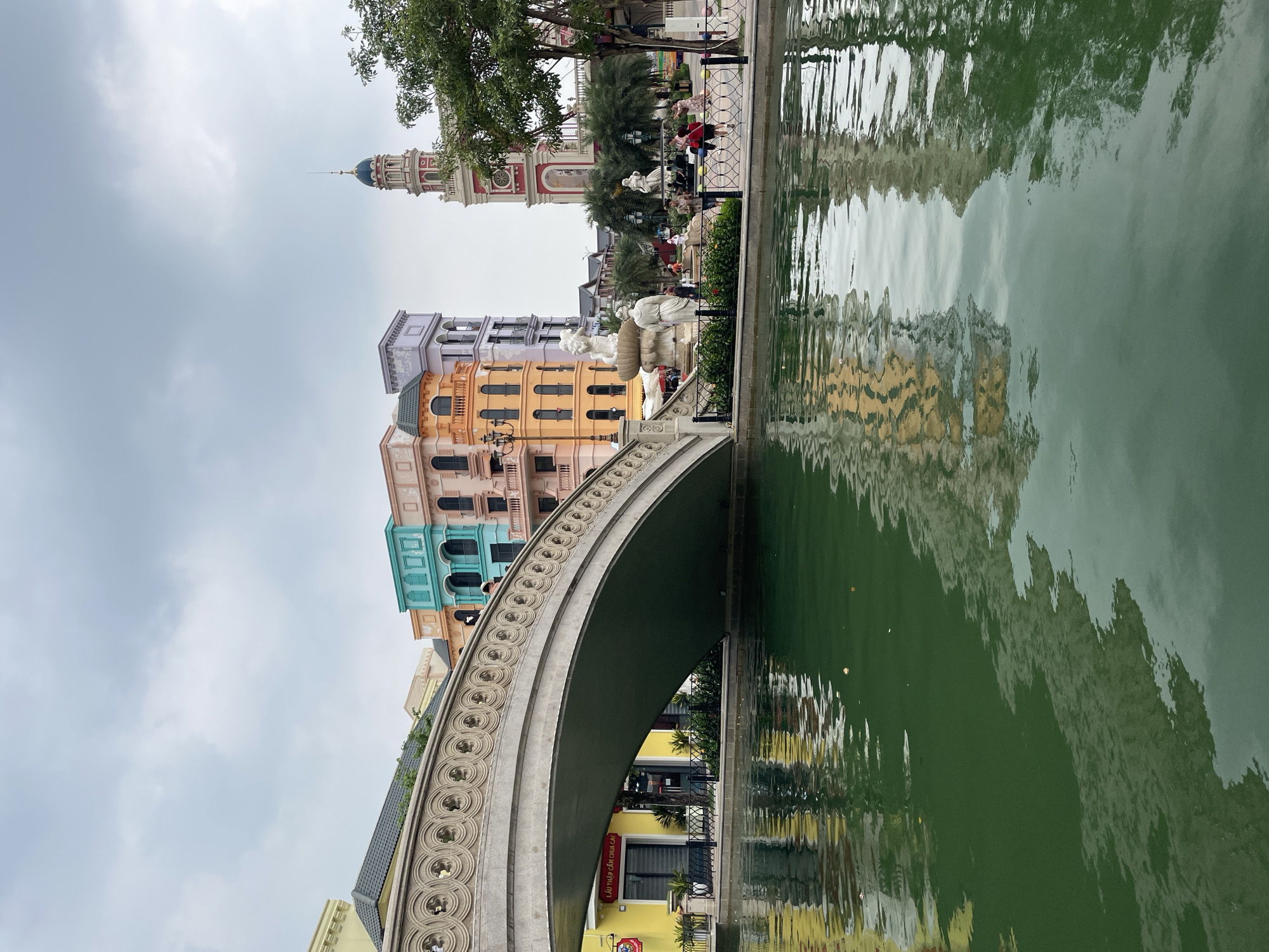 The image depicts a picturesque canal scene in the city of Nghĩa Trụ Commune, Vietnam. A stone bridge with ornate carvings spans the calm, green water, reflecting the colorful buildings on the opposite bank. These buildings feature a distinct architectural style with pastel-colored facades in shades of yellow, orange, teal, and lavender. To the right of the canal, a tall, red and white tower with a blue-domed top rises against a cloudy sky. In the middle ground, a fountain with statues is visible, and several people are strolling along the paved walkway beside the canal. Palm trees and lush greenery add to the tropical ambiance of the location.
