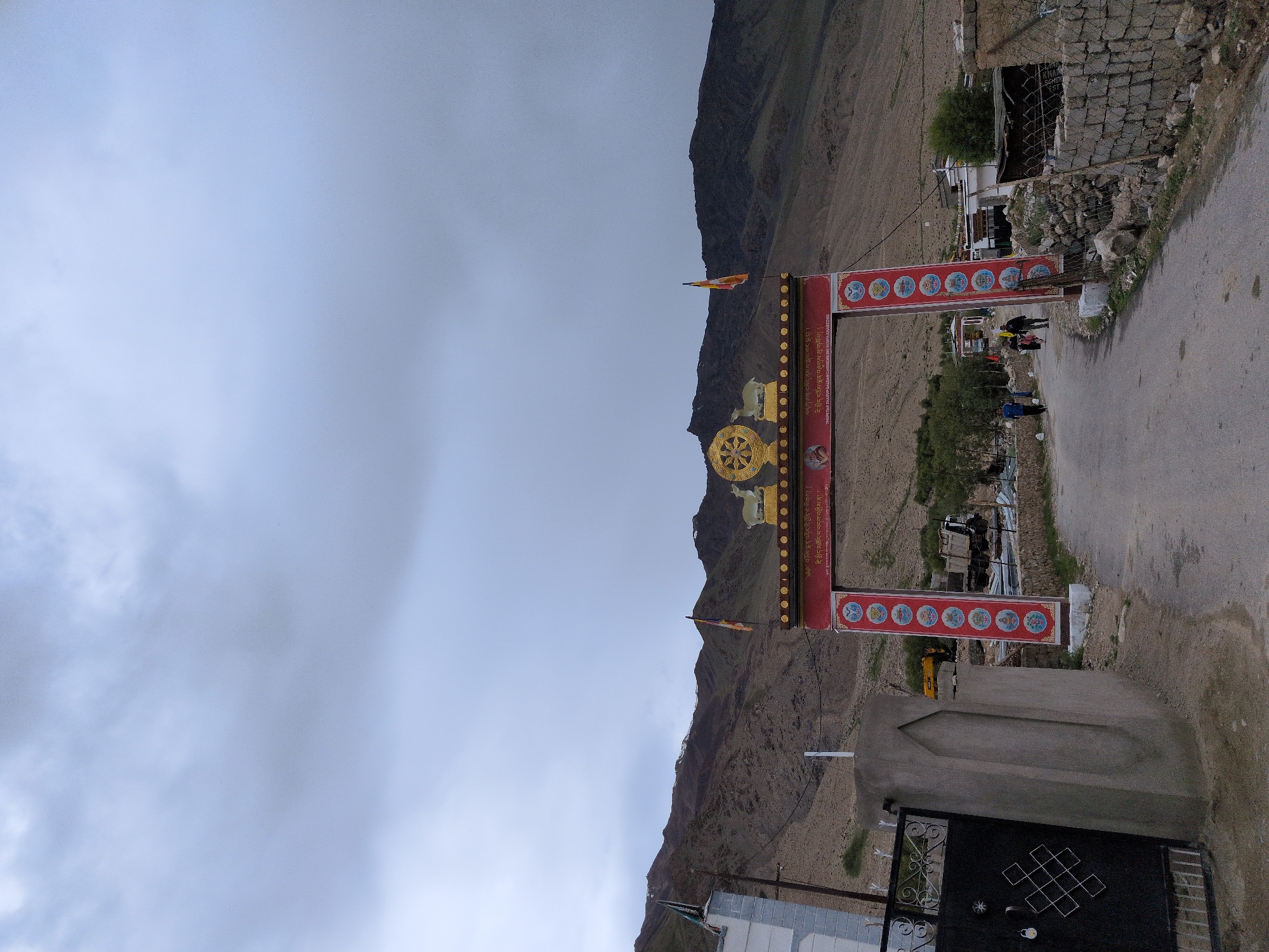 The image shows an arched gateway in Khardung, India, leading to a village. The sky is overcast with gray clouds. The gateway is ornate, with red and gold detailing, and features Buddhist iconography, including a Dharma wheel and a deer. There are also flags flying from the top of the gateway. The road leading through the gateway is paved and appears to be a village road. Several people are visible walking in the distance, and a few buildings are present on either side of the road. The surrounding landscape consists of barren, rocky hills. The overall impression is one of a remote, mountainous region with a strong Buddhist cultural presence.
