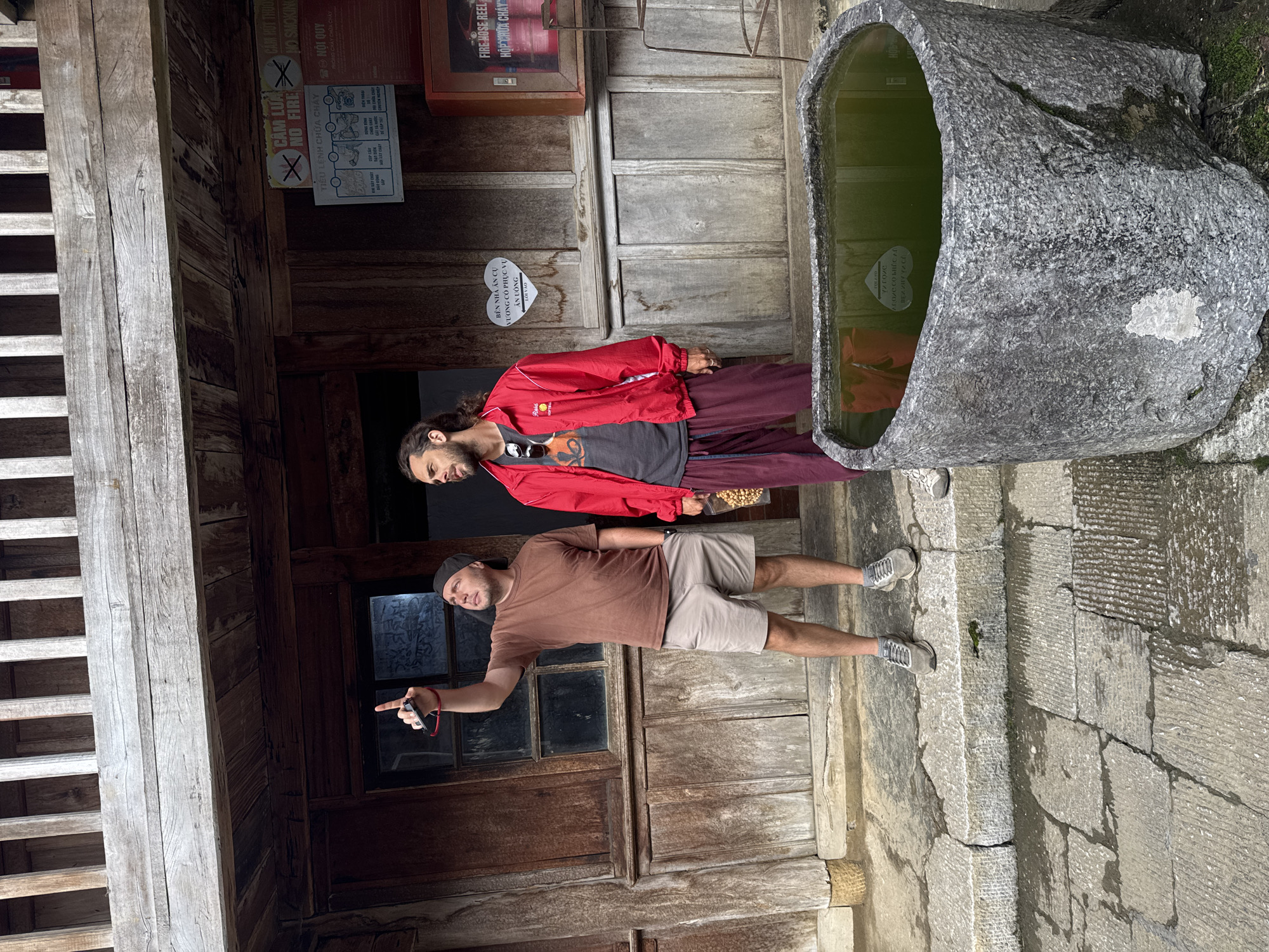 Two men stand near a stone trough filled with water in Sà Phìn Commune, Vietnam. The man on the left points towards a wooden wall with a window that has graffiti on it. Various signs, including "No Smoking" and fire safety instructions, are mounted on the wall. The other man stands beside the trough, wearing a red jacket and sunglasses. The ground is paved with stone. A wooden staircase is visible to the left.