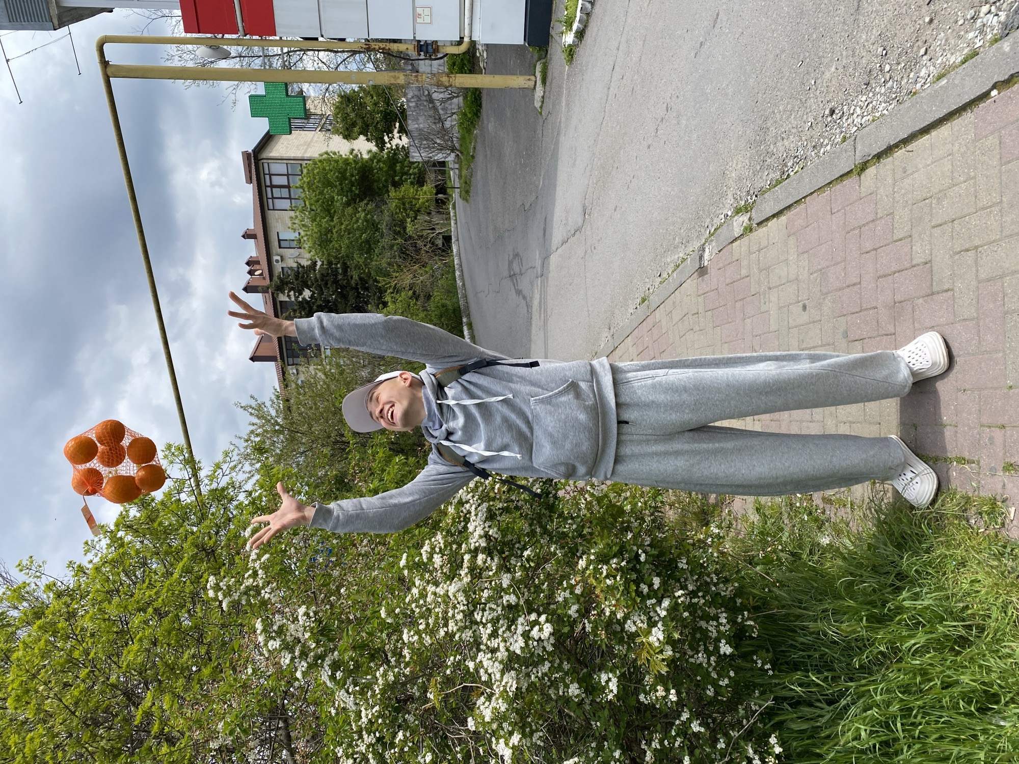 This image, taken in Gelendzhik, Russia, depicts a young person joyfully interacting with a mesh bag of oranges on a cloudy day. The individual, dressed in a grey tracksuit, a white cap, and white slip-on shoes, stands on a sidewalk with both arms raised high, hands open, and is looking upwards with a wide smile or laugh.
The mesh bag, containing several bright orange fruits (likely tangerines or oranges), appears to be suspended in the air to the left of the person. The scene is set outdoors along a street. The sidewalk features a pattern of red and beige paving bricks, bordered by an asphalt road. Lush green foliage, including bushes with numerous small white flowers and patches of green grass, lines the sidewalk.
In the background, a multi-story building with a brown roof and light-colored walls is visible. A bright green cross sign, commonly used to denote a pharmacy, is mounted on this or a nearby structure. A prominent yellow utility pipe runs horizontally across the upper part of the image, contributing to the urban landscape. The sky is overcast, indicating a cloudy daytime. While there are small labels on the fruit bag and a utility panel, no legible text is visible in the image.