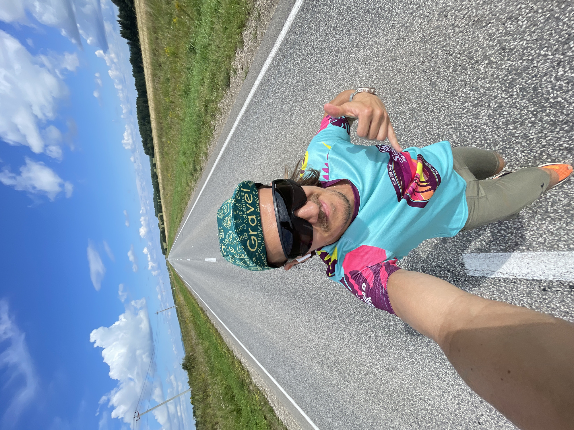 A man is taking a selfie on an empty asphalt road that stretches into the distance. He is wearing a turquoise, pink, and purple cycling jersey with text that reads "RUN FOR SUN" and "MARATHON." He also has on a green cap with "GRAVEL" printed on it, black sunglasses, and white wireless earbuds. His left arm is extended, holding the camera, and his right hand is giving a thumbs-up gesture. He is wearing khaki shorts and orange running shoes. The road is bordered by green fields and trees on either side, under a bright blue sky with scattered white clouds. The image was taken in Perevoznoe, Russia.