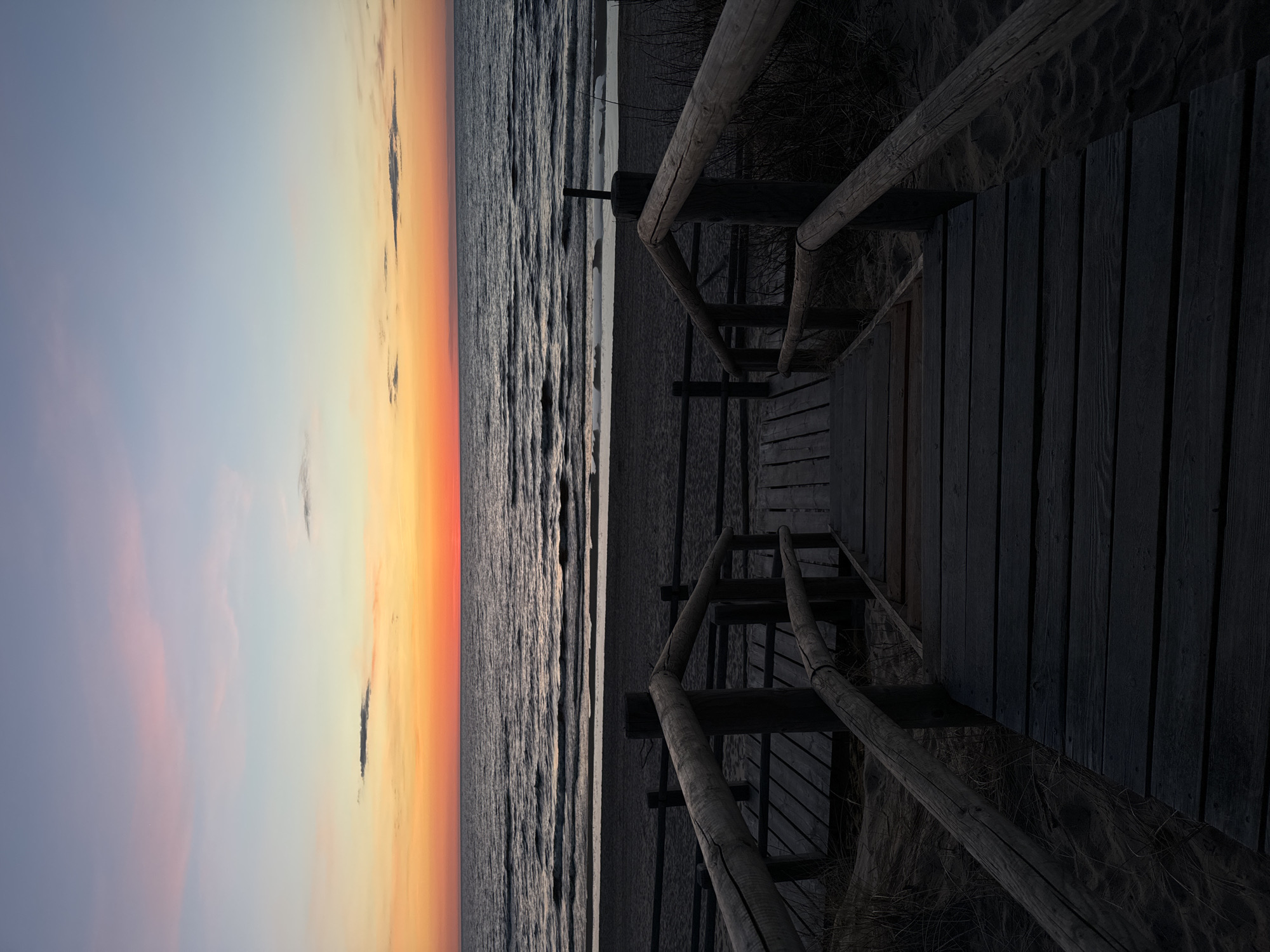The image depicts a wooden staircase leading down to a beach at sunset in Garciems, Latvia. The sky is a gradient of blue and orange, reflecting on the water. The stairs have wooden railings and are flanked by sand and vegetation. No people are visible.