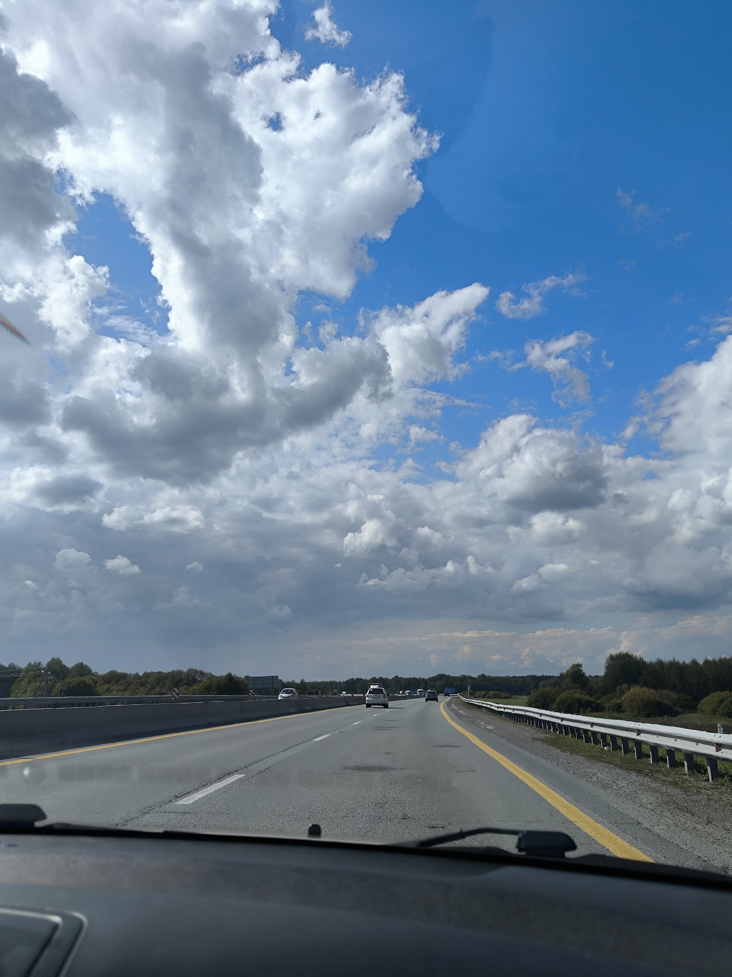 The image shows a view from inside a car driving on a highway under a partly cloudy blue sky. The highway has multiple lanes with yellow and white markings. There are cars driving in the distance. The sides of the highway are lined with guardrails and trees. The foreground shows the dashboard of the car.