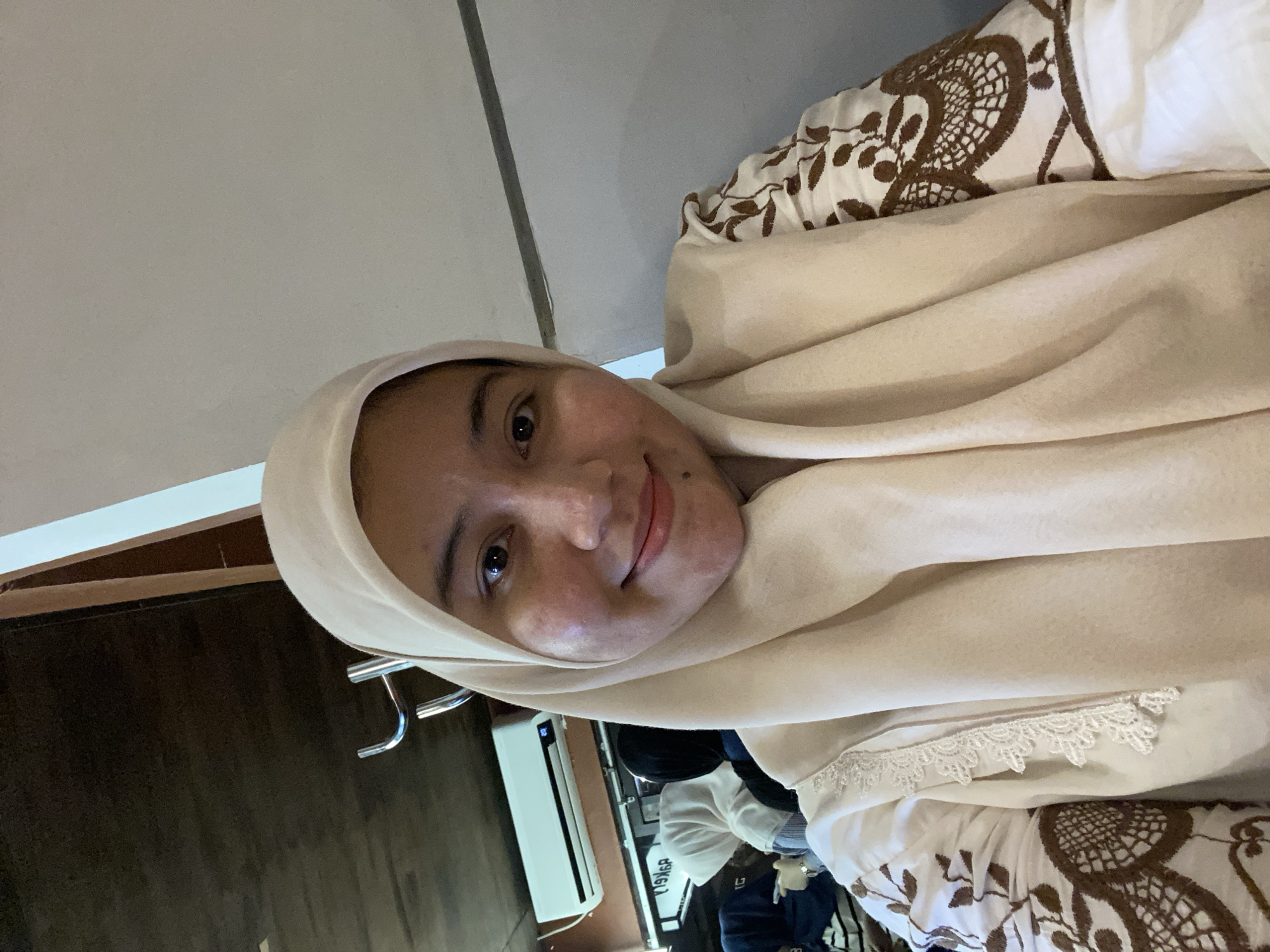 A close-up, selfie-style photograph shows a young woman wearing a light beige hijab and a matching top adorned with intricate brown embroidery on the sleeves. She has a fair complexion, brown eyes, and a slight smile. The background appears to be an indoor setting, possibly a cafe or restaurant, with dark wooden paneling on the ceiling and a white air conditioning unit mounted on the wall. Several other individuals, also wearing hijabs, are visible in the blurred background, suggesting a public gathering. The overall ambiance is casual and relaxed. This image was likely taken in Banjarbaru, Indonesia, a city known for its vibrant Muslim community and many public spaces.