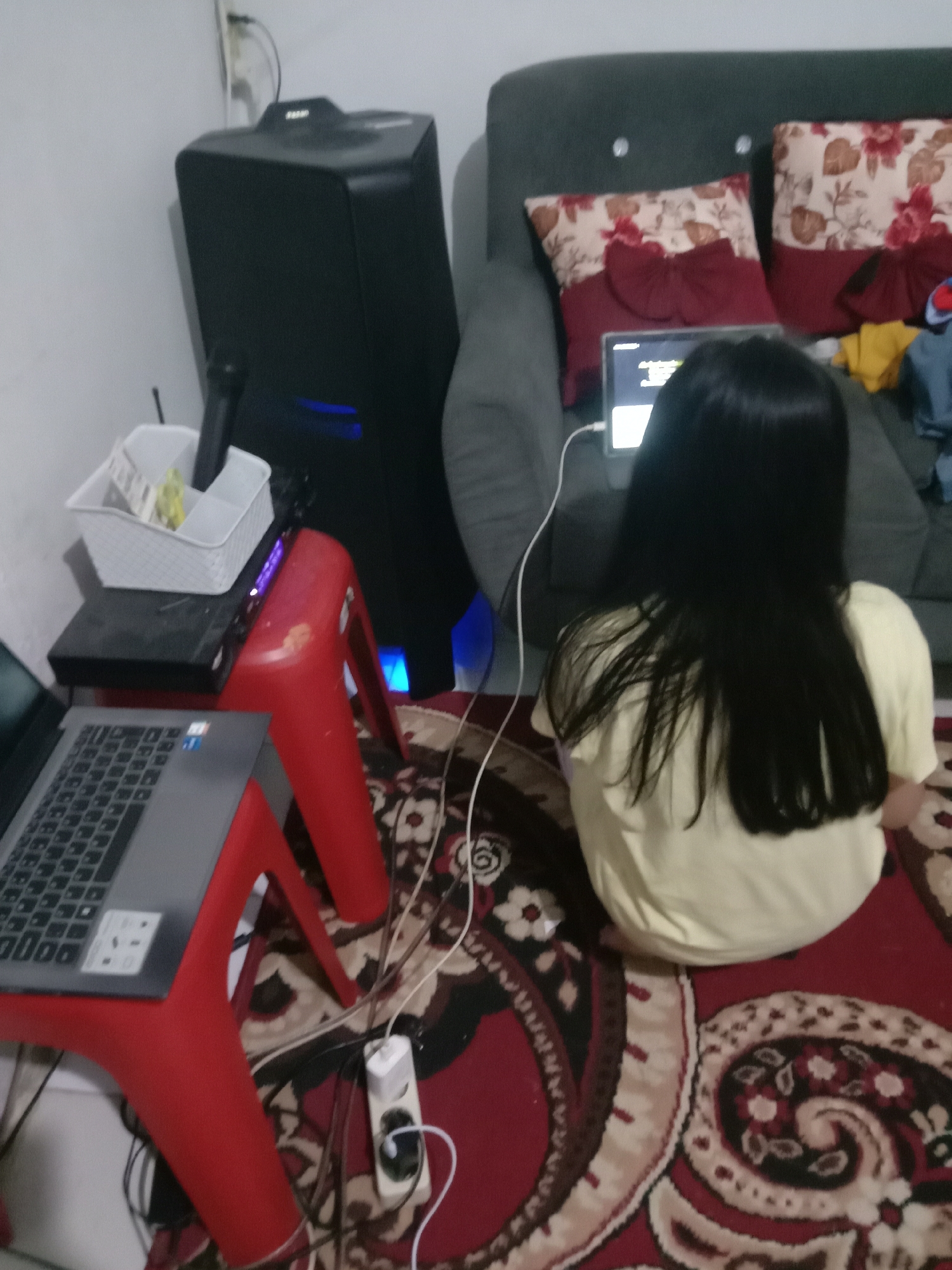 The image depicts an indoor scene, likely a living room, with a young girl sitting on the floor facing away from the camera. She is looking at a tablet or laptop placed on a couch. A large black speaker unit is positioned behind a red stool, which holds a DVD player and a white basket containing items. Another red stool, to the left, supports an open laptop. Electrical cords and a power strip are visible on the floor. The room is carpeted with a patterned rug, and the walls are white. The overall impression is one of a casual, home-based activity, possibly involving entertainment or education. The provided location of Kwandang, Indonesia, is not directly observable from the image content.