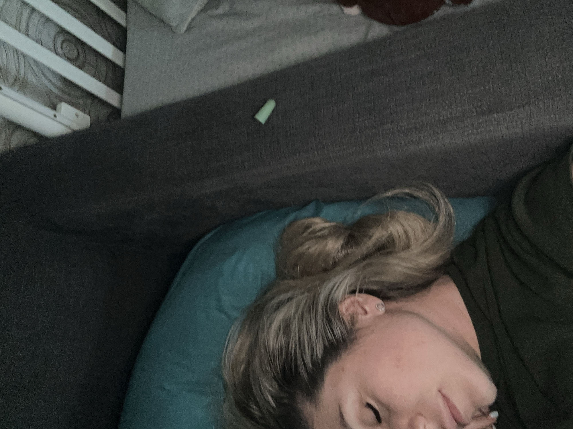 The image shows a close-up, overhead view of a blonde woman sleeping on a dark grey couch, her head resting on a teal pillow. She is wearing a dark green t-shirt and a small, clear stud earring in her left ear. Her hair is pulled back in a bun. To the right of the couch, a crib is visible with white wooden slats. A small, light green object, possibly a pill or a piece of plastic, lies on the grey fabric between the couch and the crib. The overall lighting is dim, suggesting it may be nighttime or an indoor setting with low light. There are no identifiable landmarks indicating a specific location within Novoaltaysk, Russia.