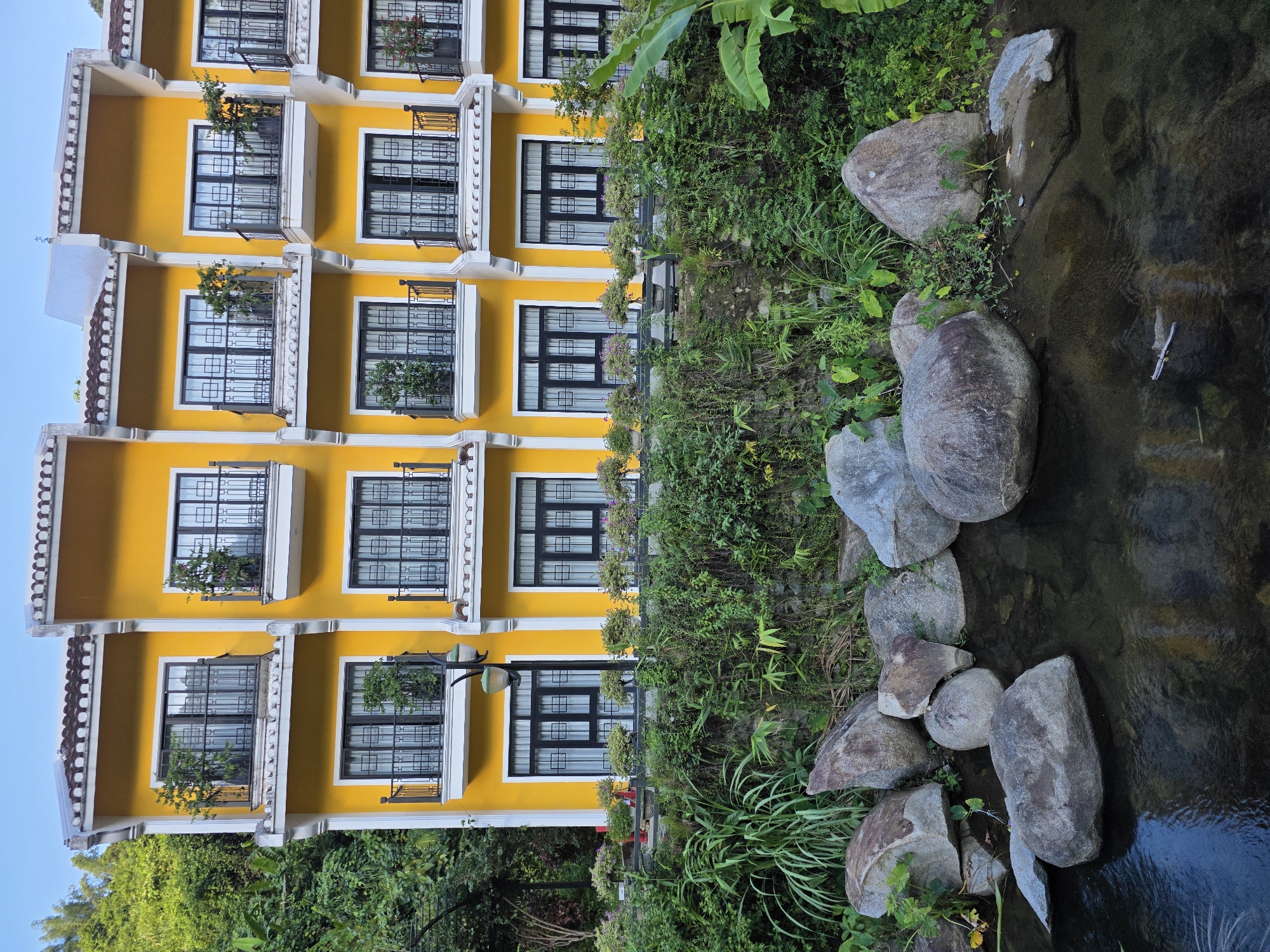 The image depicts a bright yellow, multi-story building with several balconies and windows. The building appears to be a hotel or residential complex, possibly located near a natural setting with lush green vegetation and a stream or small body of water in the foreground. Large rocks are present in and around the water. The building's architecture features white trim and dark-colored window frames. Overhead, the sky is a clear blue. No people are visible in the image.
This building appears to be the "The Blossom Resort Danang" located in Đà Nẵng, Vietnam.