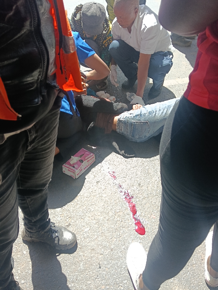 The scene depicts an outdoor emergency on a light-colored paved surface, likely a street, in Nairobi, Kenya. A person is lying supine on the ground, wearing a dark long-sleeved top and light blue denim jeans, with their head turned to the right. A distinct trail of bright red liquid, consistent with fresh blood, extends on the pavement from near the individual's head/upper body.
Two individuals are actively attending to the person on the ground. An adult male, balding and light-skinned, wearing a white short-sleeved collared shirt, dark blue trousers, and white medical gloves, is crouched on his left knee next to the supine individual's upper torso, appearing to administer aid. Another individual, wearing a blue short-sleeved top and a patterned dark and yellow fabric head covering, is crouched near the head of the person on the ground, also appearing to assist.
A pink cardboard box, possibly containing medical gloves, is located on the pavement near the supine individual's lower back. Several crumpled white cloths or gauze pads are scattered near the central figure's torso.
In the immediate foreground, to the left, a person's lower torso and legs are visible, wearing dark trousers, dark athletic shoes with white soles, and an orange item (possibly a safety vest) draped over their shoulder. To the right foreground, a person's legs are visible, clad in dark leggings and white athletic shoes. Strong shadows on the pavement indicate bright daylight conditions. Additional partial figures are visible in the background, suggesting other observers.