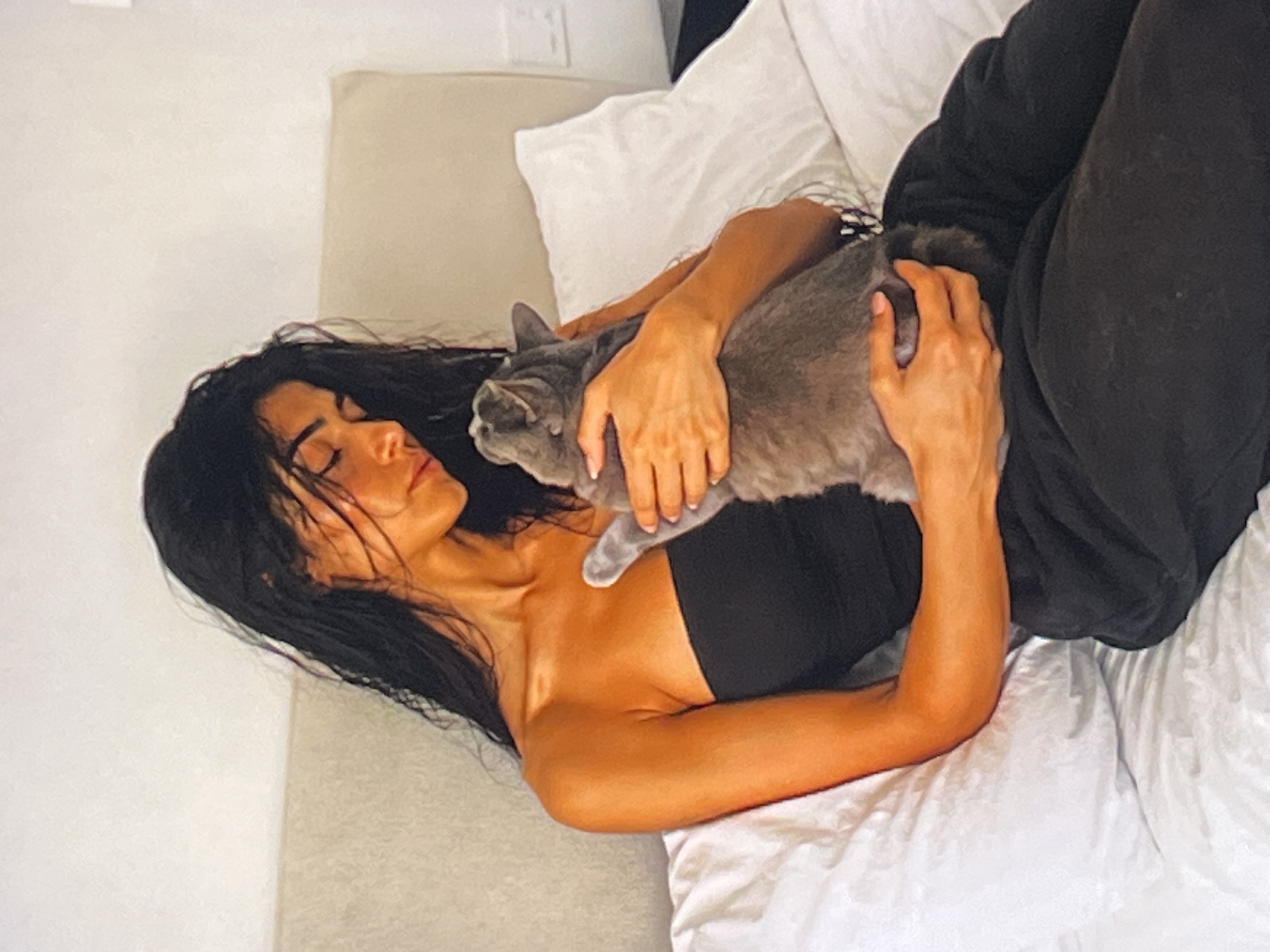 A young woman with long, dark, wet hair is sitting on a bed, holding a gray cat. She is wearing a black strapless top and dark pants. The woman is looking down at the cat, who is nestled in her arms. The bed is covered in white linens and there is a light-colored headboard behind the woman. The room appears to be a bedroom, with a light-colored wall and what appears to be a light switch on the wall. The lighting in the room is soft and warm, suggesting it might be daytime or the room is well-lit by lamps.