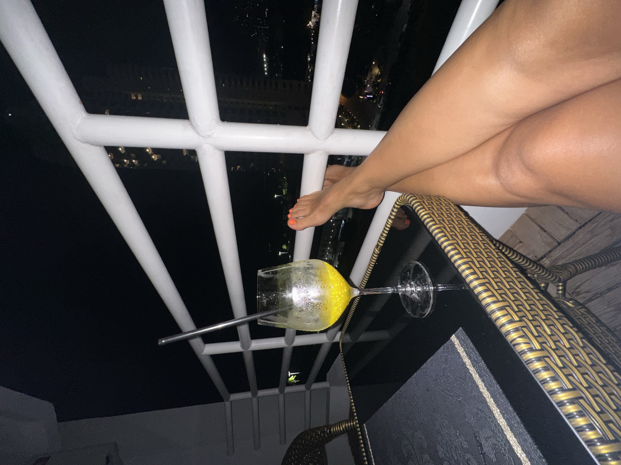 A view from a high-rise balcony at night in Pattaya City, Thailand shows crossed legs resting on a white railing. A cocktail glass with a metal straw sits on a black table with a wicker chair. Lights of buildings and street lamps are visible in the background. The legs have orange nail polish on the toes.