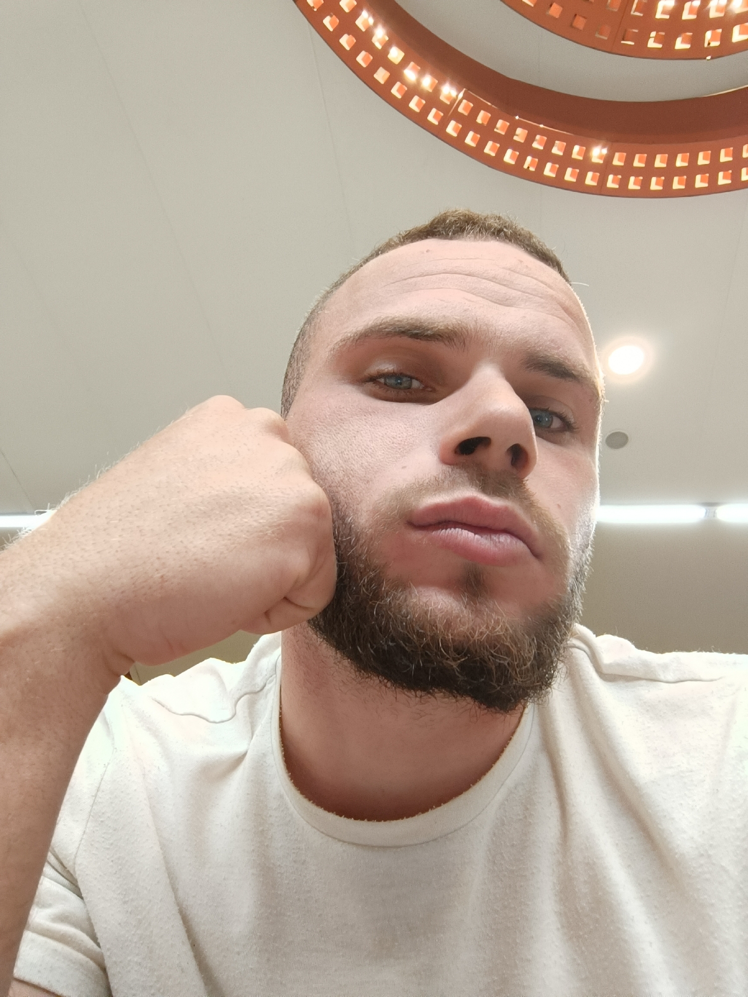 A close-up, low-angle shot of a man with fair skin and a beard. The man is wearing a white t-shirt and has his fist pressed against his cheek. The background is a light-colored ceiling with a circular, decorative light fixture.