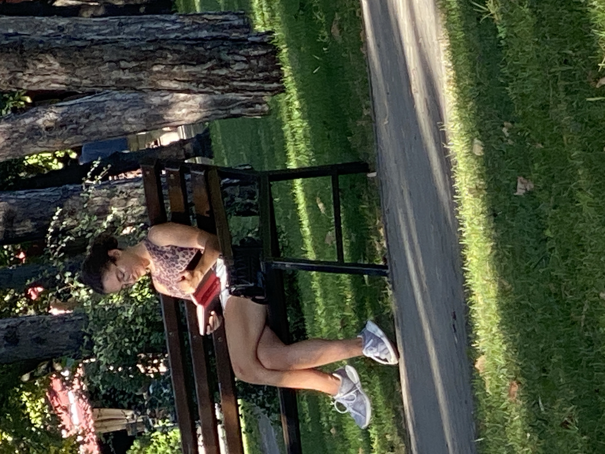 A young woman with dark hair pulled back and glasses sits on a park bench, reading a red book. She is wearing a leopard-print sleeveless top and white shorts, with grey athletic shoes. Her legs are crossed, and she is holding the book with both hands. A black purse rests on the bench next to her. The bench is made of dark wood with metal supports, and it is positioned on a paved path. The background consists of lush green grass, trees with rough bark, and sunlight filtering through the leaves, creating dappled shadows. The scene appears to be in a park in Chișinău, Moldova, given the context of the request.