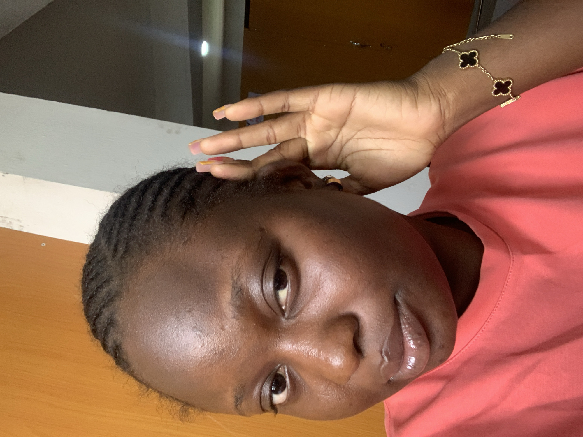 A close-up, waist-up shot captures a young Black woman indoors, likely in a residential setting. She is wearing a coral-colored t-shirt and a gold bracelet with black clover-shaped charms on her left wrist. Her hair is styled in cornrows, and she is holding her right hand up to her ear, as if listening. Her nails are painted with a pastel gradient of pink and yellow. The background features a wooden door on the left and a light-colored wall on the right, suggesting an interior space.