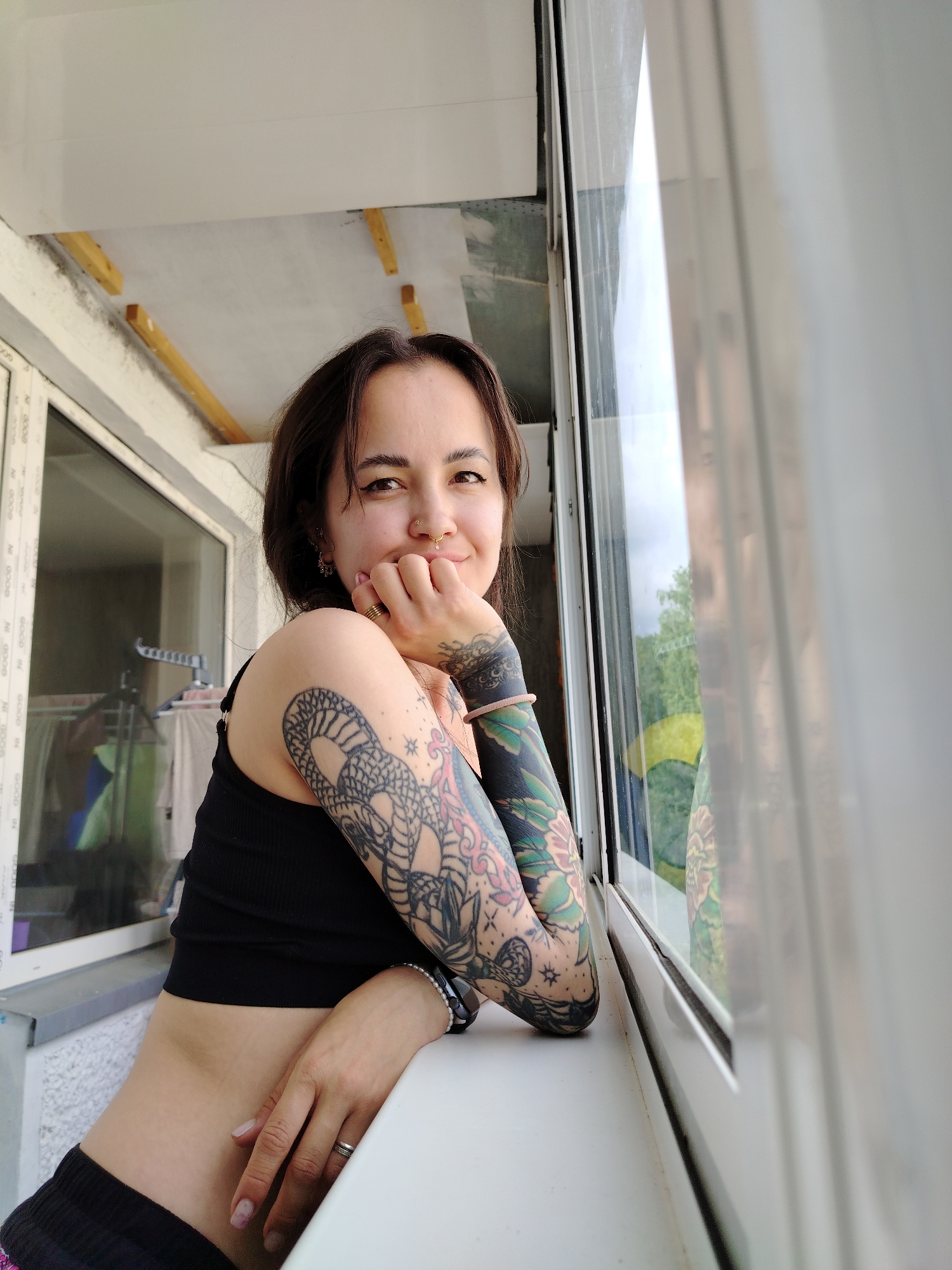 A young woman with dark hair and brown eyes smiles subtly at the camera in a medium close-up portrait. She is leaning on a white windowsill within what appears to be an enclosed balcony or sunroom, resting her left hand under her chin and her right arm on the ledge. She wears a black cropped top and dark bottoms.
Notable features include extensive, colorful sleeve tattoos on both arms, showcasing intricate designs that resemble traditional Japanese motifs such as snakes, flowers, and patterns. She has multiple facial piercings, including a septum ring and a nostril ring, and wears rings on her fingers and bracelets on her right wrist.
The background consists of white UPVC window frames and glass panes. Through the glass, green trees and bright daylight are visible, suggesting a clear day. Reflections in the window also show parts of what appears to be a clothes drying rack. Above, unfinished ceiling elements with exposed wooden beams are visible. Faint black text, reading "N 9000 IN GOOD..." or similar, is visible on the left window frame. The scene conveys a relaxed, comfortable atmosphere, likely in a residential setting in Noviy, Russia.